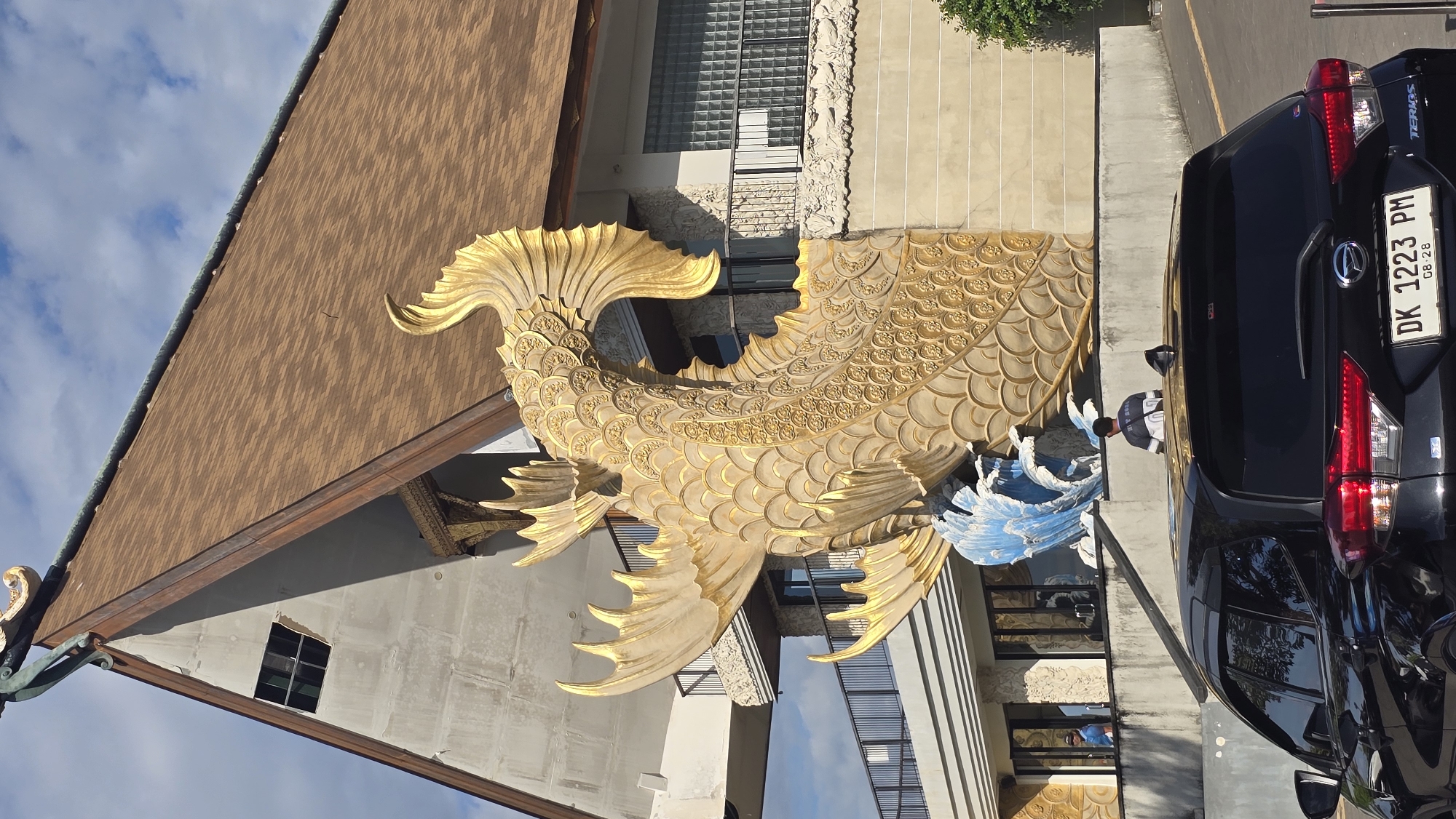The image displays an outdoor scene featuring a large, ornate golden fish sculpture as a prominent focal point. The sculpture, intricately detailed with scales and flowing fins, appears to be an architectural or decorative element. Below the golden fish are blue, wave-like structures.
To the left of the sculpture is a building with a brown tiled roof and light-colored walls, featuring both regular and glass block windows. A black Daihatsu Terios SUV is parked to the right of the sculpture. The car's rear is visible, displaying the Indonesian license plate "DK 1223 PM" with "08-28" below it. "DK" is the regional code for Bali, aligning with the Denpasar location context.
Two individuals are present: one person in a striped shirt and dark pants is standing near the sculpture and the car, while another person is faintly visible as a reflection in a nearby window. The lighting suggests it is daytime, with bright sunlight illuminating the golden sculpture and casting shadows. The sky is clear with scattered clouds. The scene gives the impression of a decorative entrance or outdoor area, possibly for a commercial building or residence in Bali.