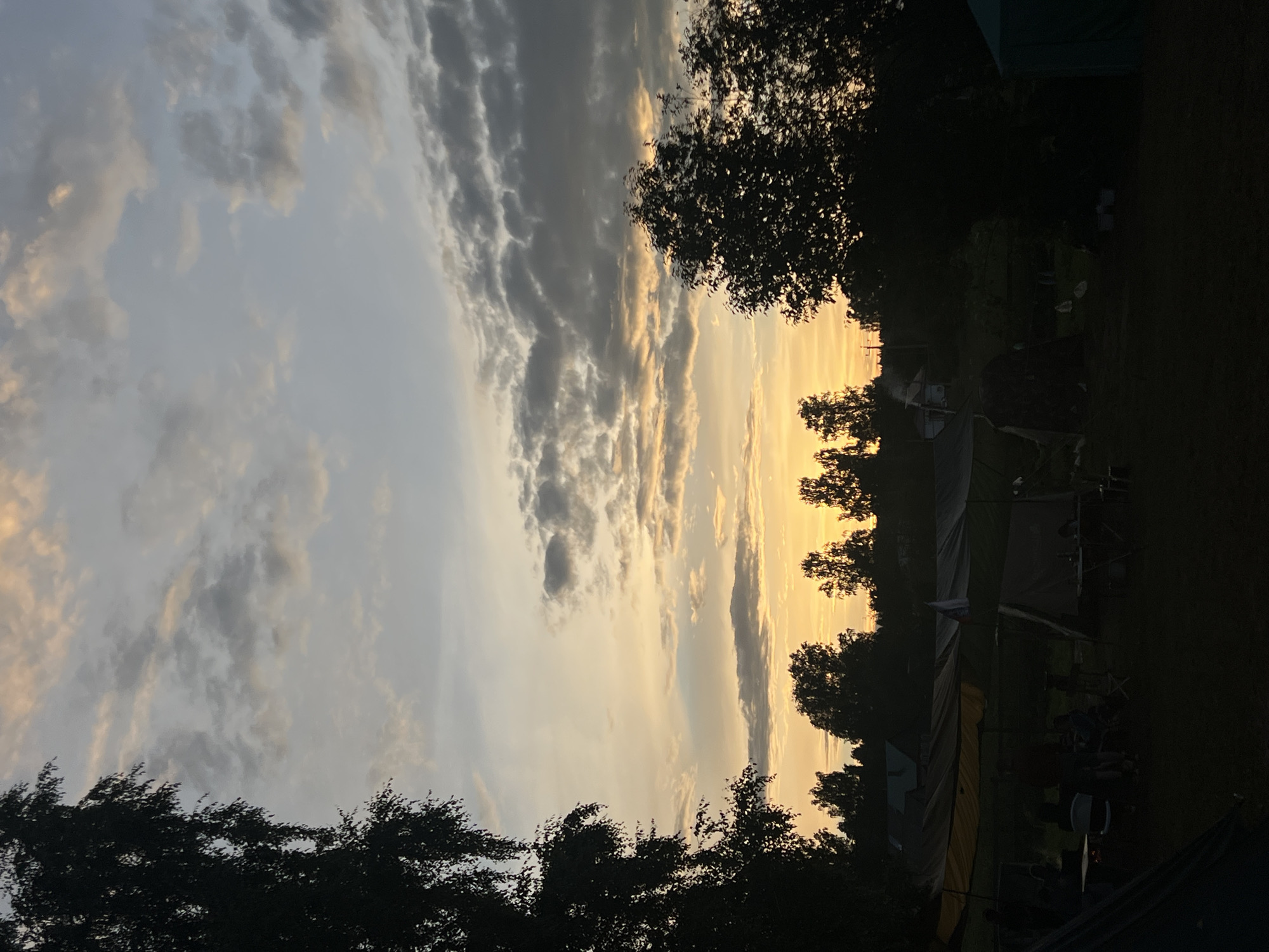 This media file depicts a scene at dusk in Sergino, Russia, likely at a campsite. The sky is filled with dramatic clouds, transitioning from a pale grey on the left to a warm, golden-yellow glow on the right, indicating the setting sun. Silhouetted trees frame the sky on the left and right edges of the image. In the foreground and mid-ground, several tents are set up, suggesting a camping or outdoor gathering. Some figures, appearing as dark silhouettes, are visible near the tents, indicating the presence of people. There is also camping equipment such as chairs and tables scattered around. The overall mood is peaceful and serene, characteristic of an evening outdoors. The time of day is clearly sunset, with the low angle of the sun creating long shadows and a warm color palette in the sky. There is no visible text in the image.