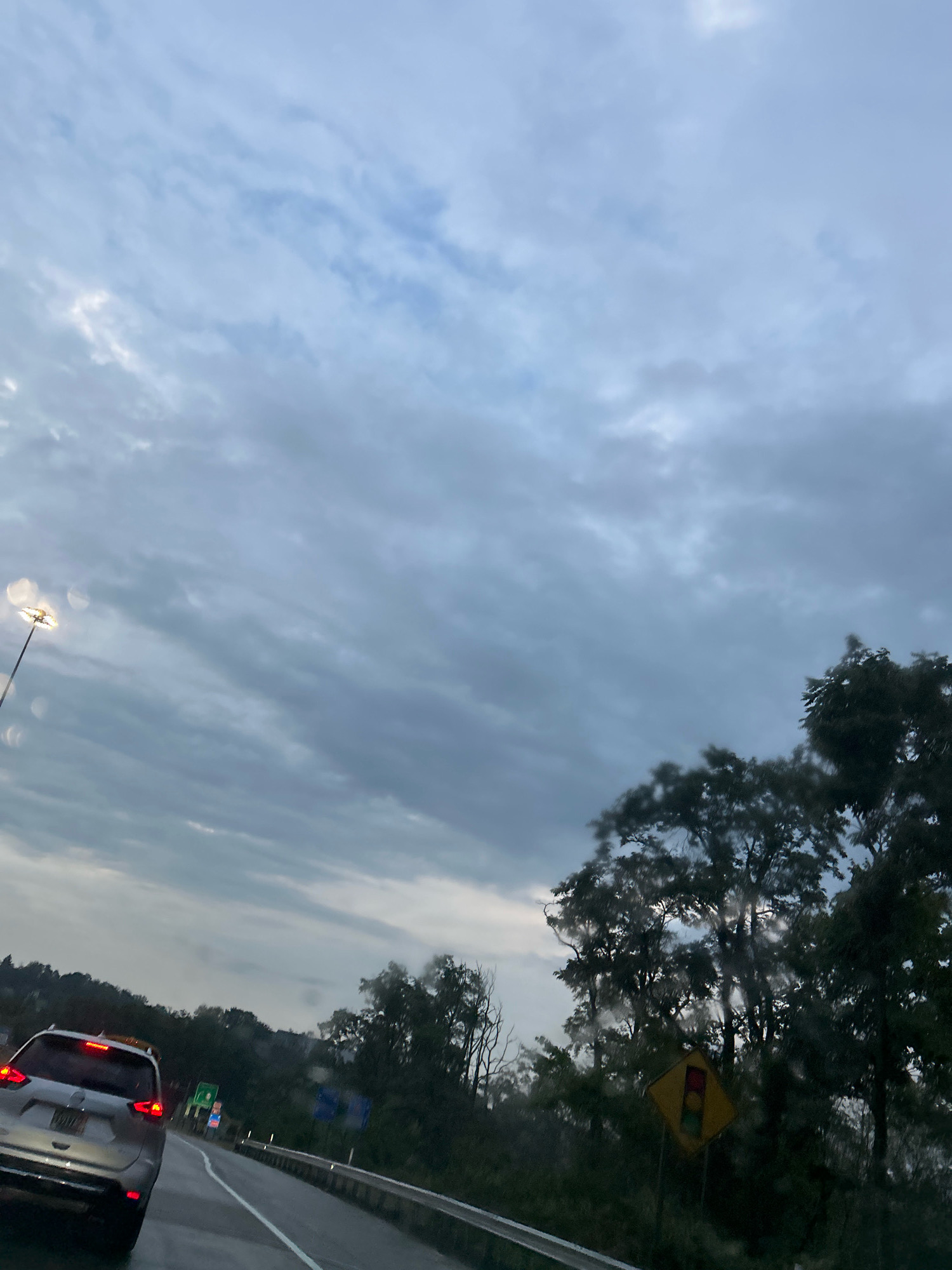 A view from a vehicle traveling on a highway in Robinson Township, United States of America, shows a cloudy sky overhead with light illuminating from a street lamp on the left. The road ahead curves to the right, bordered by trees and a guardrail. A silver SUV with its brake lights on is in the foreground, and further ahead, traffic signs are visible. The scene appears to be set in the late afternoon or early evening.