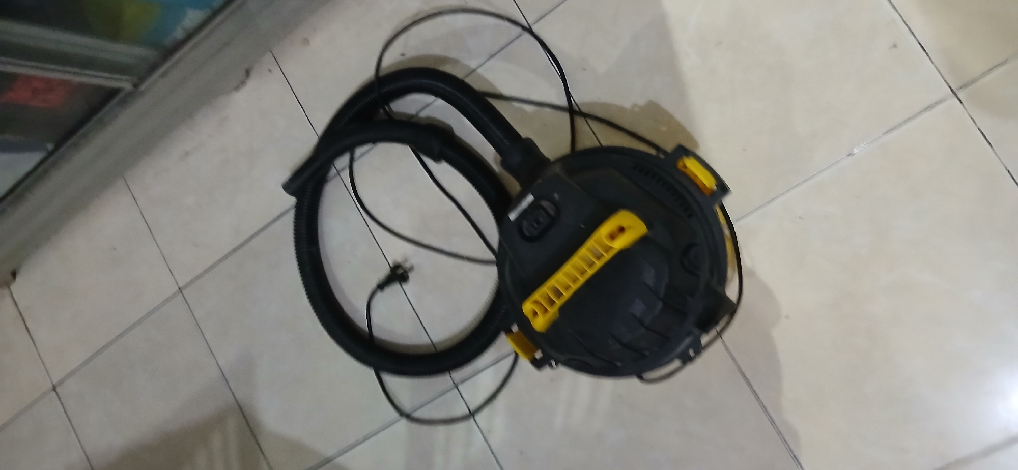 A black and yellow shop vac with a black corrugated hose and power cord is resting on a tiled floor. The shop vac is circular in shape with a yellow handle on top and yellow latches on the side. The tiled floor is composed of light-colored square tiles with dark grout lines. The scene appears to be indoors, possibly in a storage area or garage, in Samarinda, Indonesia. The background shows the edge of a cabinet with glass doors. There are no people visible in the image.