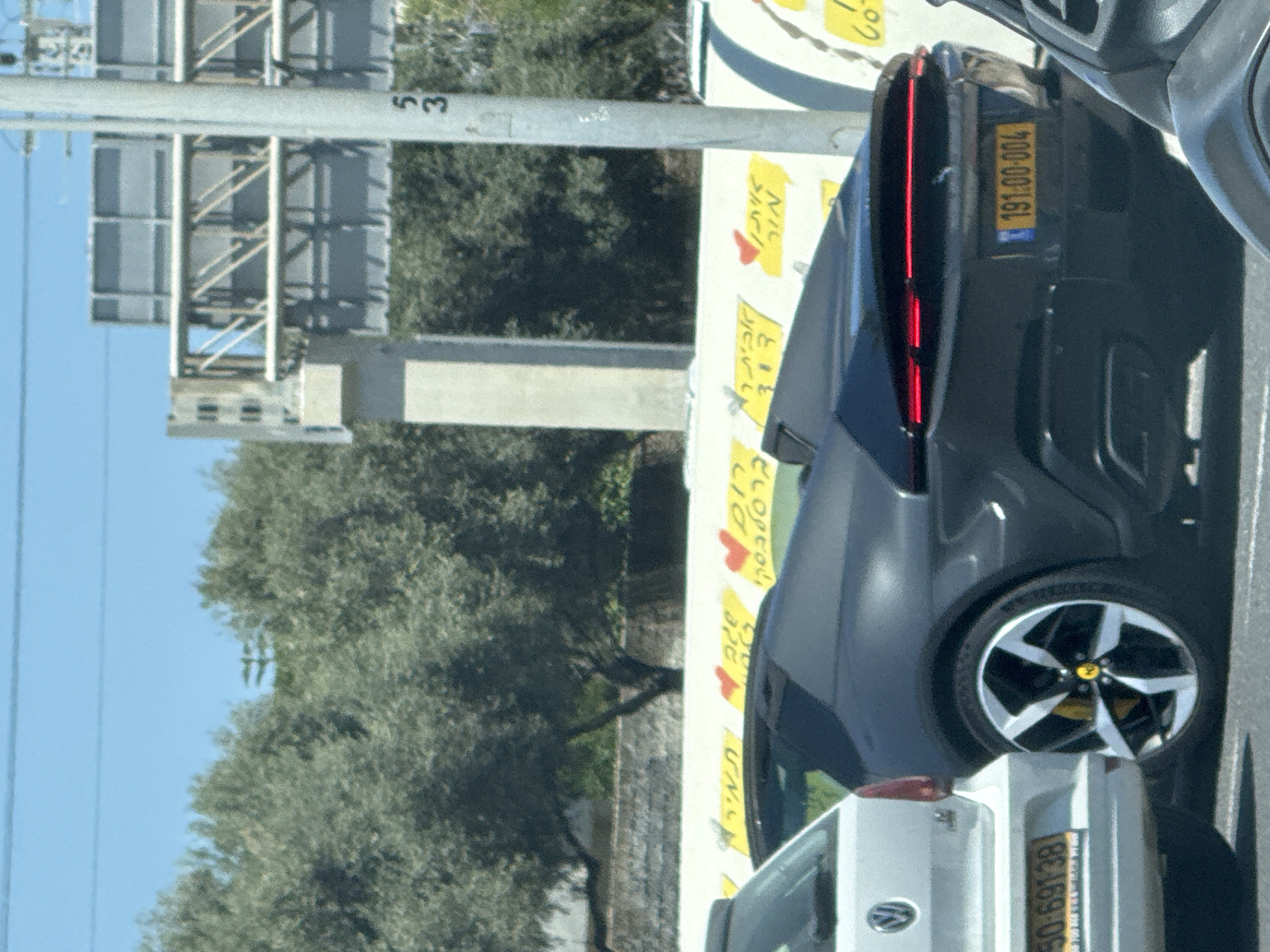 The image captures a scene in what appears to be Herzliya, Israel. A gray car, identified by its license plate "191:00-004", is parked next to a white vehicle with the plate "50:691-38". Roadside advertising is visible with yellow signs bearing Hebrew text and heart shapes. A highway structure and the road marker "53" can be seen in the background, alongside a tree.