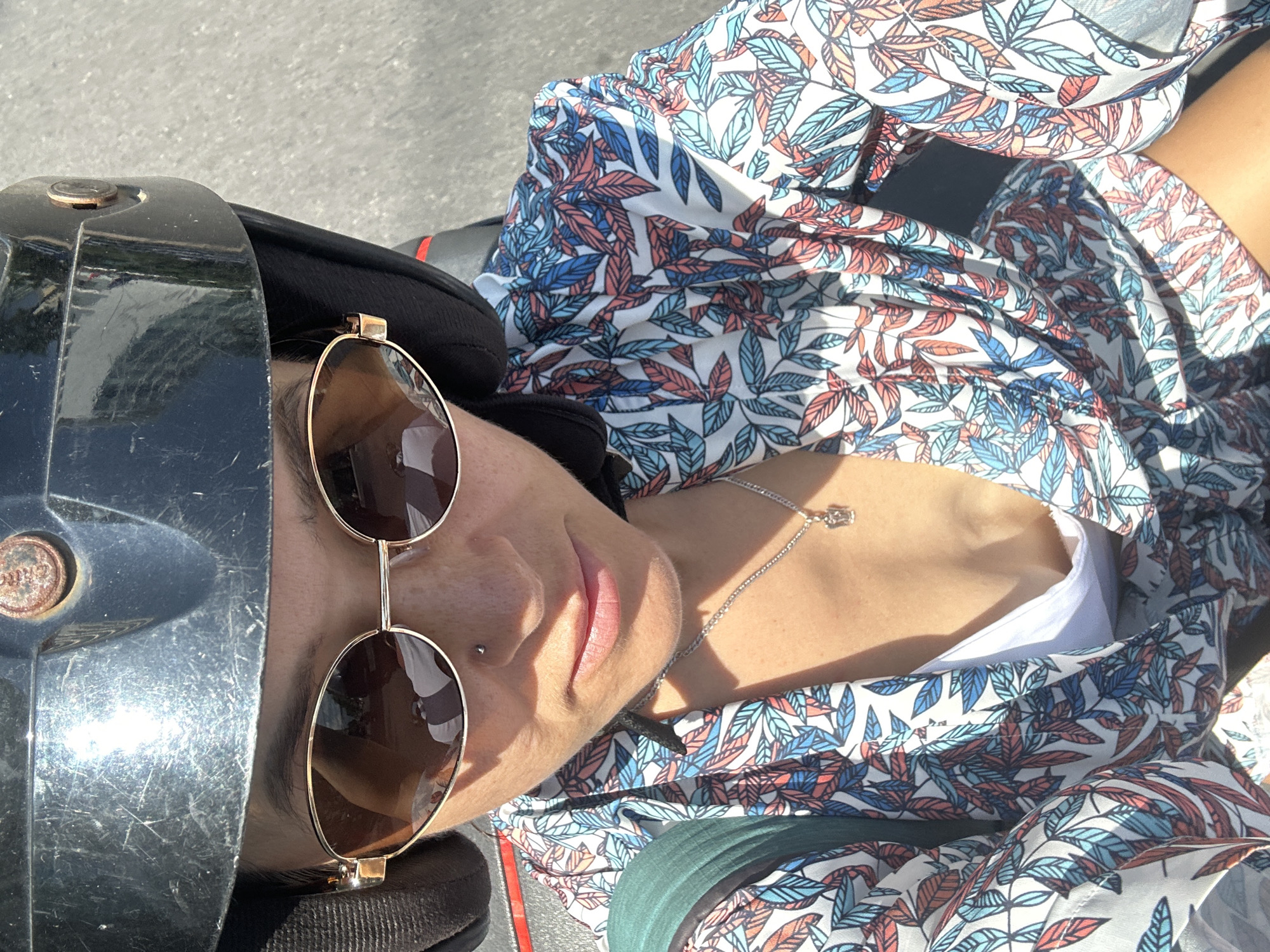 A medium shot shows a woman from the chest up, wearing a black helmet and oversized sunglasses. Her hair is pulled back, and she has a nose ring. She is wearing a white top and a colorful, patterned shirt with leaves in blue, red, and white. A silver necklace with a small pendant is visible. The background is an asphalt road, suggesting she is on a motorcycle or scooter. The image appears to be taken on a sunny day, with shadows cast by the subject. The location is identified as Nha Trang Ward, Vietnam.