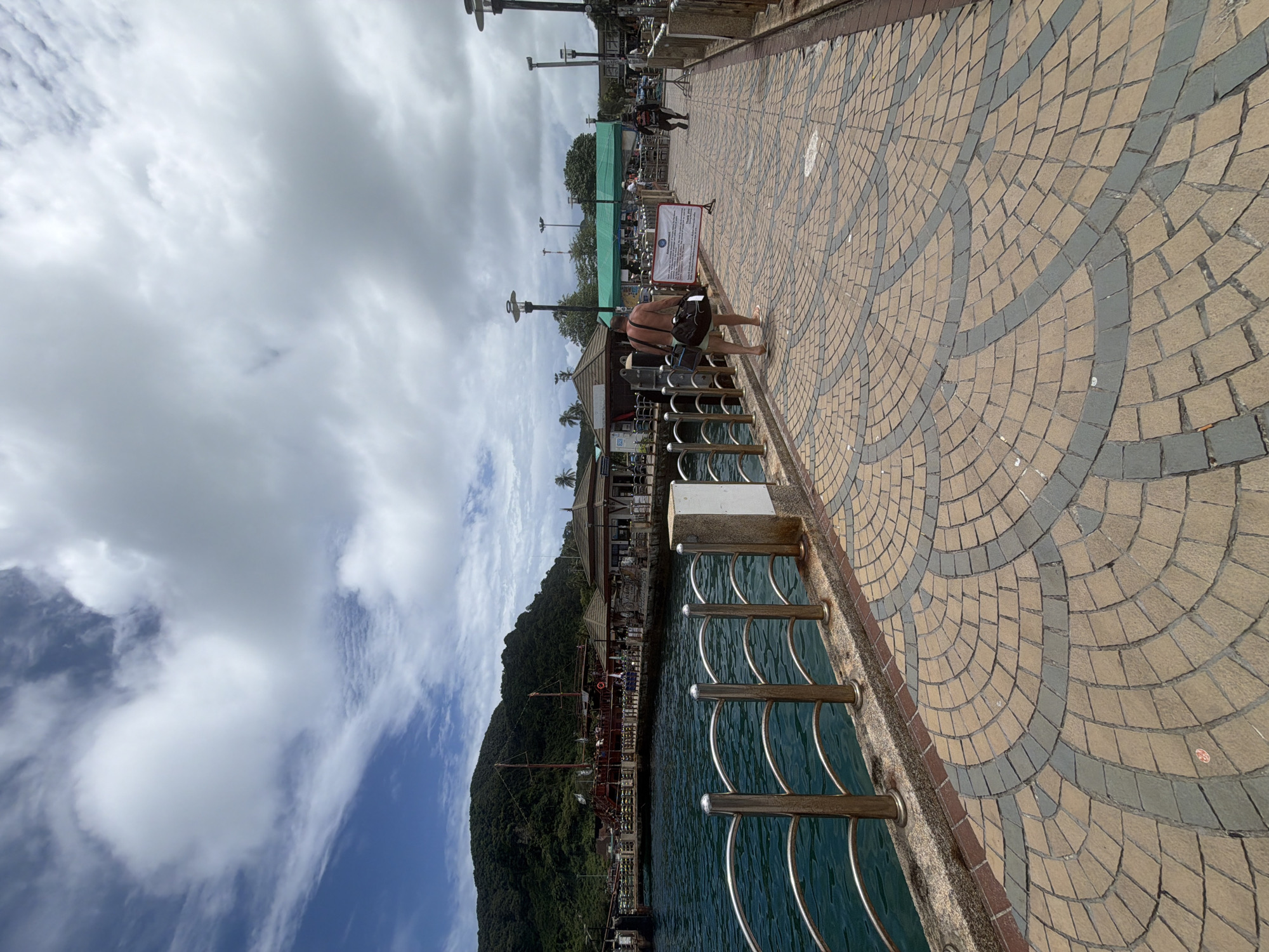 The image depicts a waterfront promenade in Ao Nang, Thailand, likely a pier or walkway leading to a ferry terminal or boat docking area. The foreground features a cobblestone pathway with a repeating circular pattern of light brown and gray tiles. A metal railing with horizontal bars runs along the left side, bordering the teal-colored water.
In the midground, a man walks away from the camera, carrying a dark bag over his shoulder and wearing shorts and flip-flops. Further along the pathway, another person in dark clothing is visible. Several people are also seen near structures in the background.
The background reveals a large, traditional-looking wooden boat with multiple masts docked in the water. Adjacent to the boat are low-rise buildings with distinctive, pointed roofs, suggesting commercial establishments or waiting areas. Beyond these structures, a lush green, mountainous landscape rises, characteristic of the region. The sky is partially cloudy with white clouds against a blue background. A few lampposts are visible along the pathway. A sign with text is positioned on the right side of the walkway.