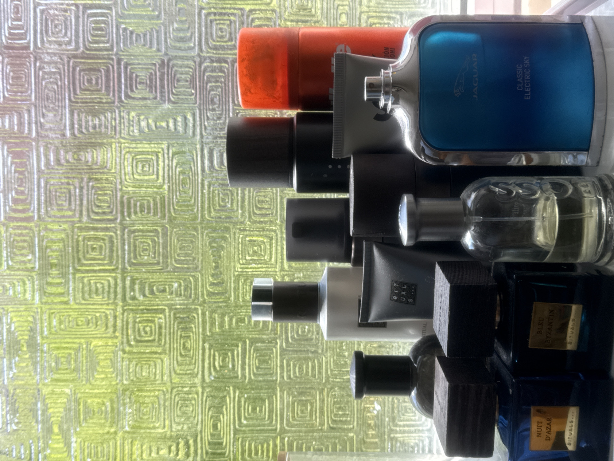 A still life image showcases a collection of men's grooming products and perfumes arranged on a white surface in front of a textured, green-tinted glass window. The window features a repeating pattern of concentric squares. The foreground displays two rectangular blue glass perfume bottles, each with a dark wooden cap and a gold label. The left bottle is labeled "NUIT D'AZAR" and "RITUALS...", while the right is labeled "BLEU BYZANTIN" and "RITUALS...". To the right of these is a clear glass perfume bottle with a silver spray top, containing a light yellow liquid, and further to the right is a large, blue, glass perfume bottle with a silver rim and cap, featuring the Jaguar logo and the text "CLASSIC ELECTRIC SKY". Behind these, a variety of other bottles and tubes are visible, including a white bottle with a black cap, a gray tube, several dark, cylindrical containers, and a bright orange aerosol can. The lighting suggests it is daytime, with natural light filtering through the patterned glass. The scene appears to be indoors, possibly on a windowsill or shelf in a bathroom or dressing area. The arrangement of items suggests they are personal care products. The location is identified as Oeiras, Portugal.
