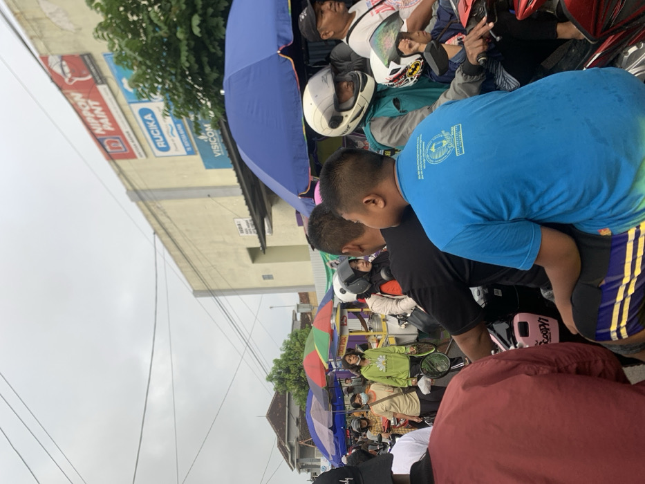 The scene is a crowded street in Kecamatan Lawang, Indonesia, located at coordinates -7.848 latitude, 112.699 longitude. The street is lined with shops, including one with a large sign that says "NIPPON PAINT". Several people are crowded around the street. A man in a blue t-shirt is standing in the foreground, while several other people are visible in the background. Some of the people are holding umbrellas. A motorbike is parked in the foreground. The sky is cloudy. The people appear to be waiting in line for something. They are interacting with each other by talking and looking at each other. The overall scene is crowded and busy.