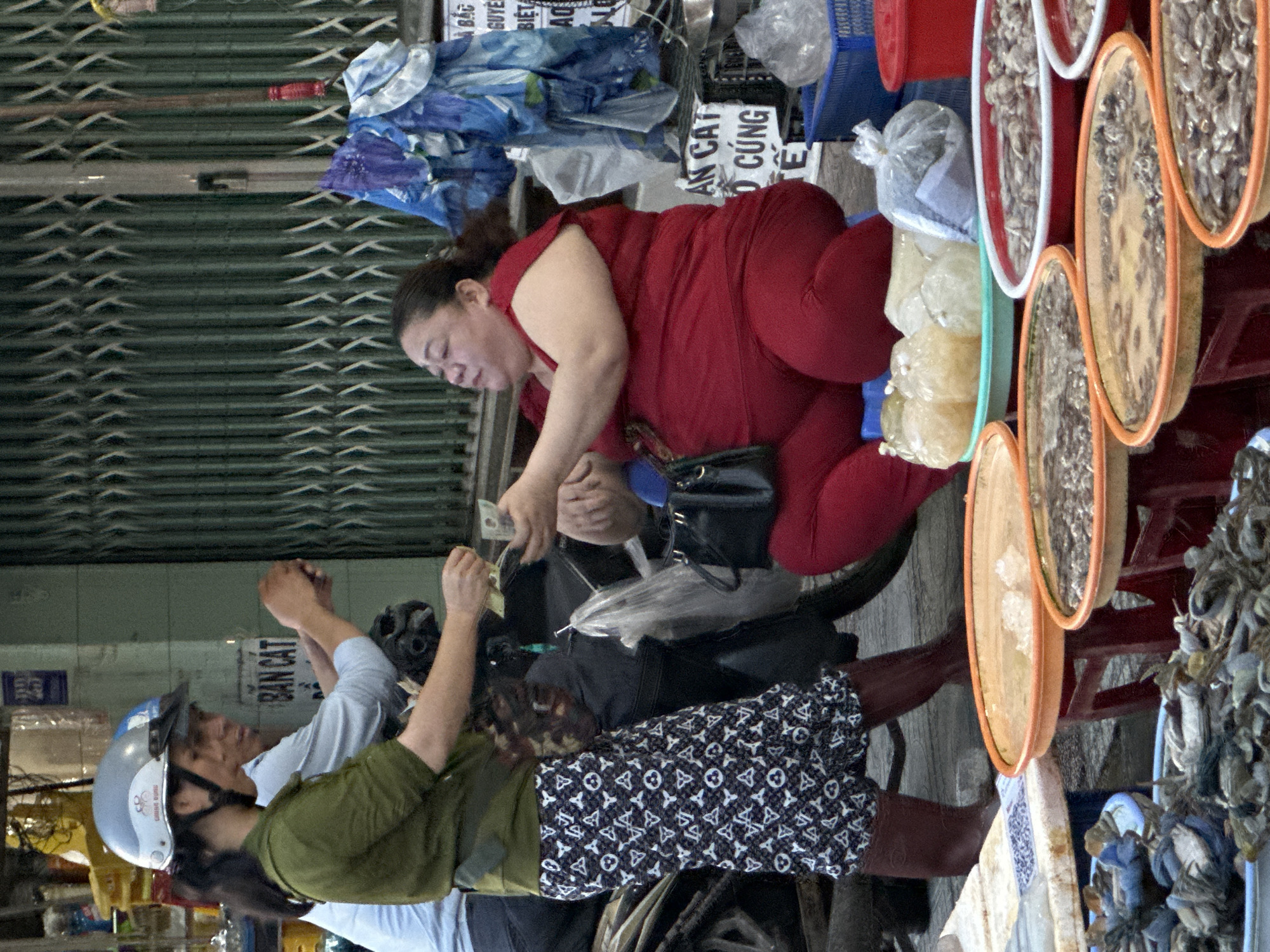 At an outdoor market in Nam Nha Trang Ward, Vietnam, two people are engaged in a transaction. On the left, a person wearing a green shirt and a patterned skirt, with a white helmet on their head, is handing money to someone on the right. The person on the right is wearing a bright red top and red pants. They are seated and appear to be a vendor, with various items displayed on tables in front of them, including seafood in bowls and bags of goods. A motorcycle is parked behind the two individuals. The background shows a green metal gate and a wall. The lighting suggests it is daytime.