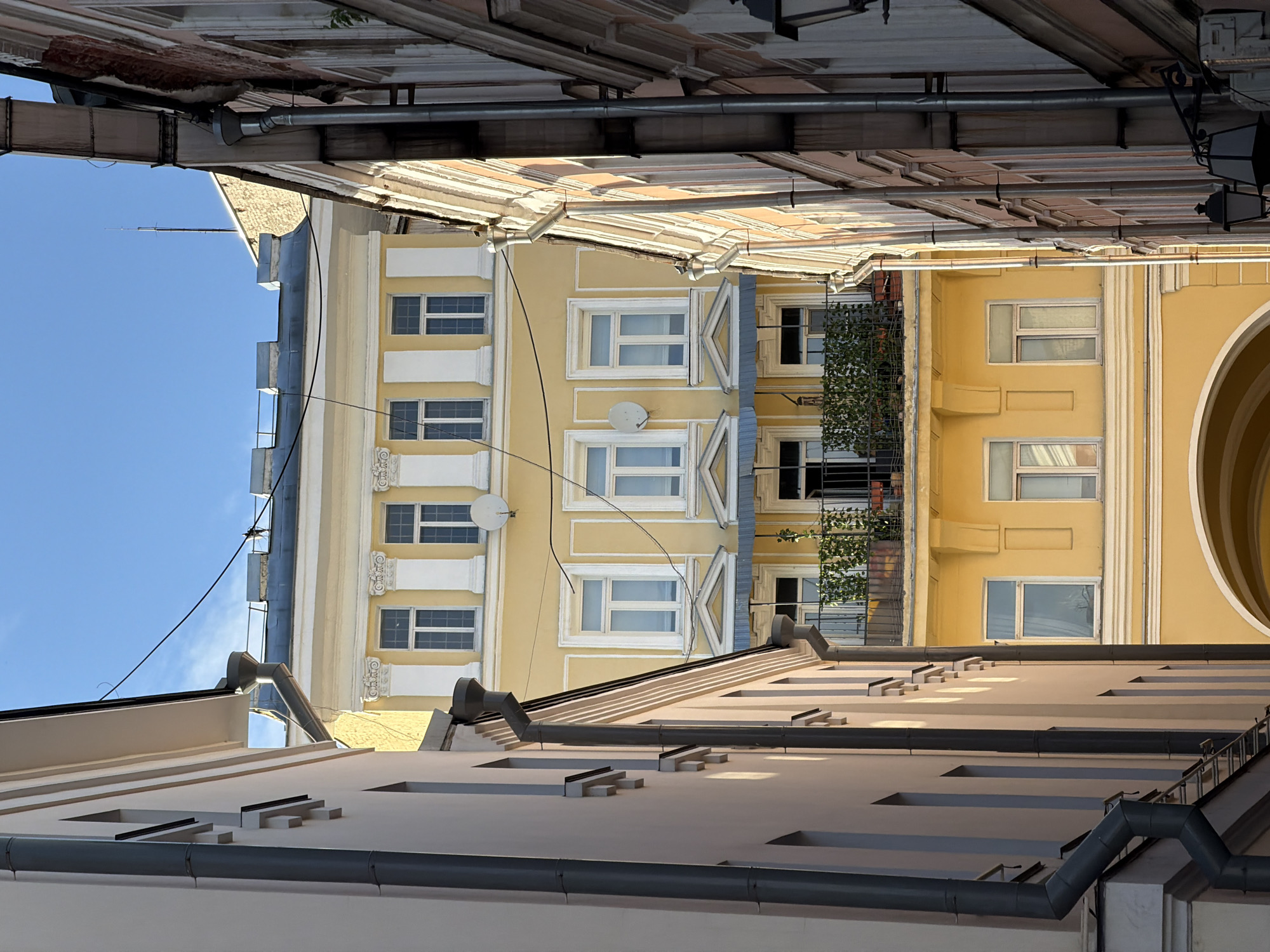 The image is a low-angle view of buildings in Moscow, Russia, seen from within a narrow space between them. The sky is visible in the upper left corner, partially obscured by the edges of the roofs above. A yellow building with white-framed windows and decorative elements stands prominently in the center. A satellite dish is visible on the side of the yellow building. A building with a balcony covered in greenery is seen to the right of the yellow building. Below, a light-colored building with horizontal architectural features and a gray gutter is in the foreground. The overall impression is that of a dense urban environment.
