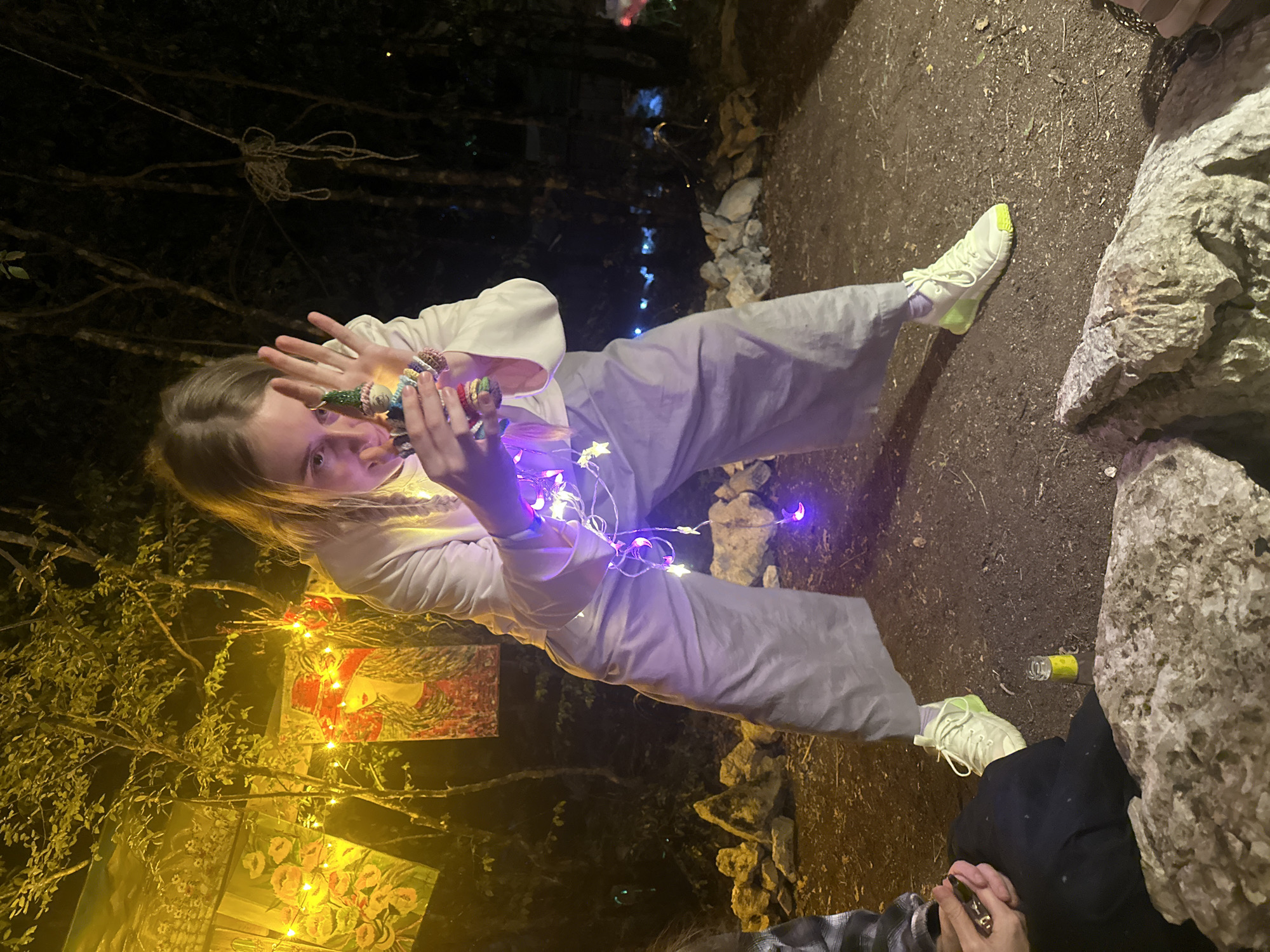 The image depicts a person holding a small, colorful, knitted doll with a pointed hat, adorned with purple star-shaped lights. The person is wearing a white long-sleeved top and wide, light gray pants, with white sneakers featuring neon yellow accents. They are standing outdoors in what appears to be a dimly lit area with trees in the background. The ground is covered in dirt and small rocks. To the left, there are two large, light-colored rocks in the foreground. Behind the person, there are tapestries or paintings hanging, illuminated by yellow lights. A string of purple star-shaped lights is draped around the person and the doll. A small glass bottle with a yellow cap is on the ground near the person's feet. The scene suggests an outdoor gathering or event at night.