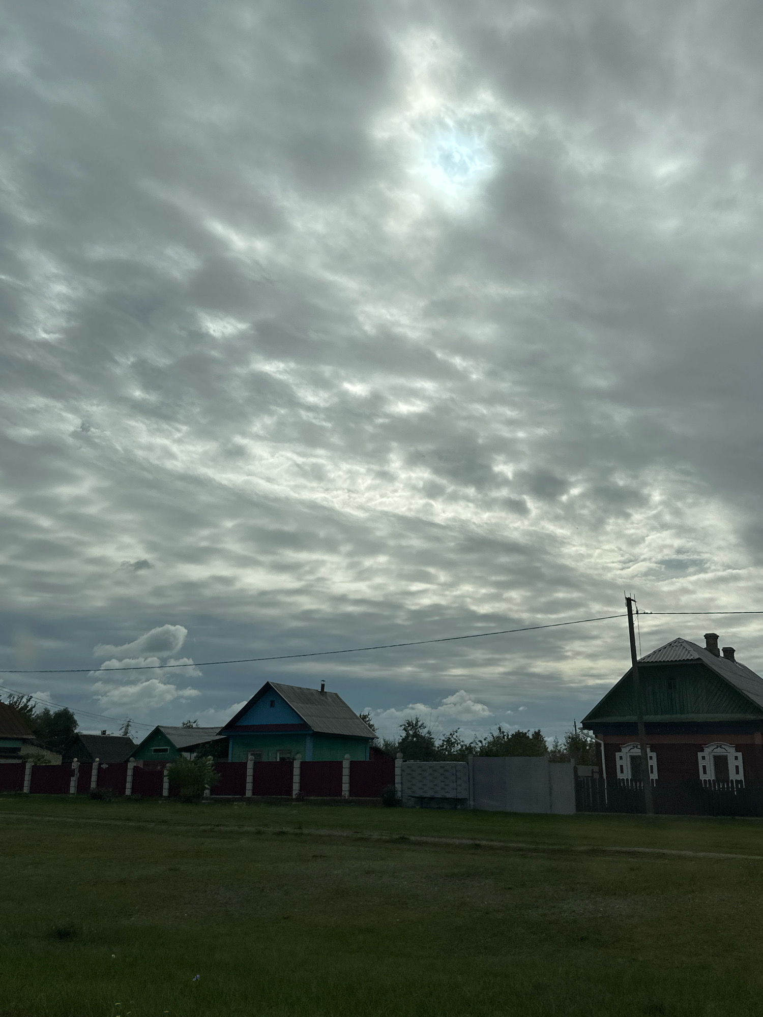 The image depicts a rural scene in the Parytsky Selsoviet, Belarus, under a dramatic, overcast sky with a bright sun breaking through in the upper center. A grassy field occupies the foreground, leading to a street lined with residential houses and fences. On the right, a dark red house with a green roof features decorative white window frames and two chimneys. To its left, a lighter-colored house with a turquoise upper section and a green roof is bordered by a dark red fence. Further to the left, other houses and fences are visible, suggesting a village or a collection of rural dwellings. A utility pole with wires stands near the red house, and a light grey concrete fence is situated between some of the properties. The overall impression is of a cloudy day in a Belarusian village.