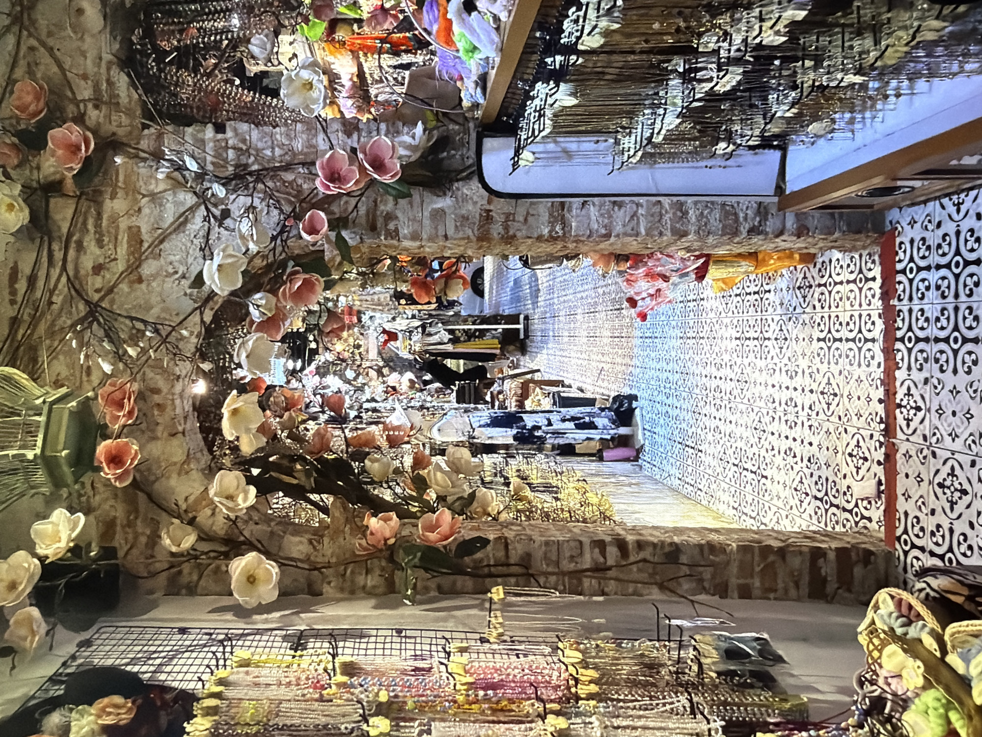 This image captures the interior of what appears to be a vibrant and elaborately decorated boutique or shop, viewed from a slightly elevated perspective. No people are visible in the scene.
The setting is characterized by a rich, eclectic aesthetic. In the foreground, on the left, there's a display area featuring various jewelry or craft items arranged on wire grids, showcasing an array of colorful bracelets or necklaces. Above and around this area, numerous artificial flowers in shades of white and pink, attached to branches, cascade downwards, creating a lush, organic feel. A decorative, light-colored birdcage is also visible on the far left.
The midground reveals more of these floral arrangements, some entwined around what looks like an archway or decorative wall section made of bricks or stone, further enhancing the whimsical atmosphere.
Towards the background and right side of the image, the store extends into a longer space. The floor is covered with striking patterned tiles, primarily in black and white, creating a visually engaging surface. Shelves and racks filled with merchandise line the walls, and a long display counter or table, reflecting bright light, runs parallel to the tiled floor, showcasing more items. Overhead, various decorative elements and lighting fixtures are suspended from the ceiling. Baskets filled with colorful textiles or items are placed on the floor towards the right edge.
The overall impression is that of a bustling, artfully arranged retail space, possibly a gift shop, craft store, or fashion boutique, designed to attract customers with its intricate decor and diverse offerings. The lighting suggests an indoor setting, likely during operating hours. There is no visible text.