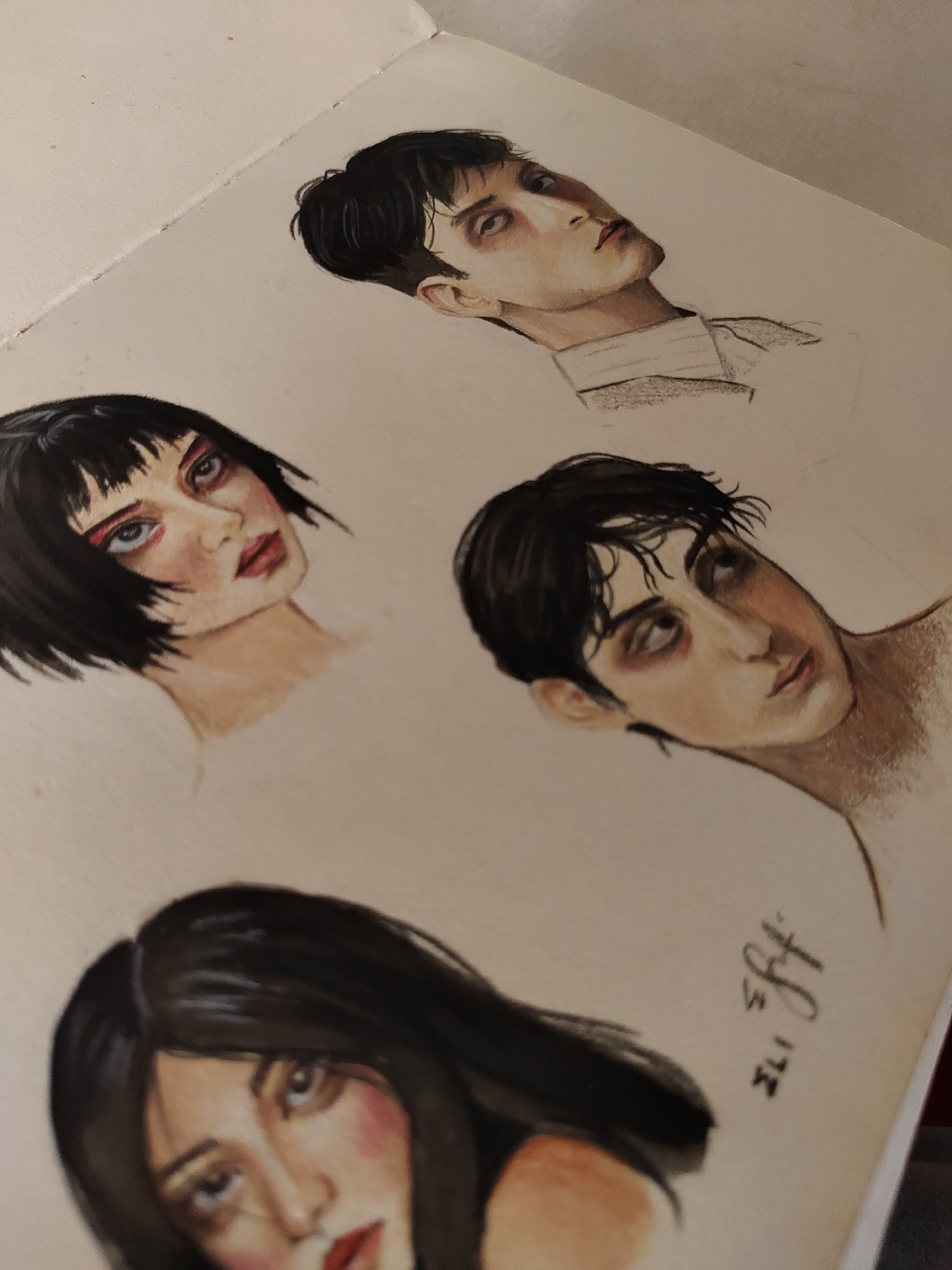 The image displays a page from an open sketchbook or art pad, featuring four distinct portrait drawings or studies. The medium appears to be colored pencils or pastels, with a stylized, expressive aesthetic. The paper is light-colored, possibly off-white or cream, showing a visible fold or binding line in the upper left.
**Subjects:**
- **Upper-right portrait:** A male figure with dark, neatly styled hair, looking upwards and to the left. He has prominent cheekbones, subtly colored lips, and an intense, slightly melancholic expression. A collared shirt is lightly sketched around his neck.
- **Mid-left portrait:** A female figure with a short, dark bob haircut and bangs. Her gaze is direct, with large eyes heavily accentuated by red or pink makeup and dark shadowing. Her lips are full and red, and her neck is lightly sketched.
- **Mid-right portrait:** A male figure with dark, somewhat tousled hair, head tilted, looking upwards and to the right. His eyes show prominent dark circles or reddish shadowing, giving him a fatigued or dramatic appearance. His skin is pale with a blush on the cheek.
- **Bottom portrait:** Partially visible, out of focus, showing the upper part of a female face with long, dark hair. She has soft blush on her cheeks and red lips, with her gaze directed slightly downwards.
**Activity:** The image captures a collection of finished or in-progress portrait drawings, showcasing an artist's work or practice.
**Notable details:** The drawings emphasize expressive facial features and dramatic eye makeup or shadowing. The overall impression is one of intense, moody, or character-driven studies. Lighting appears soft and diffused, highlighting the artwork.
**Visible text:** A stylized signature or initials, reading "ILI SFJH" or similar, is present vertically on the right side of the page, between the middle-right and bottom portraits.