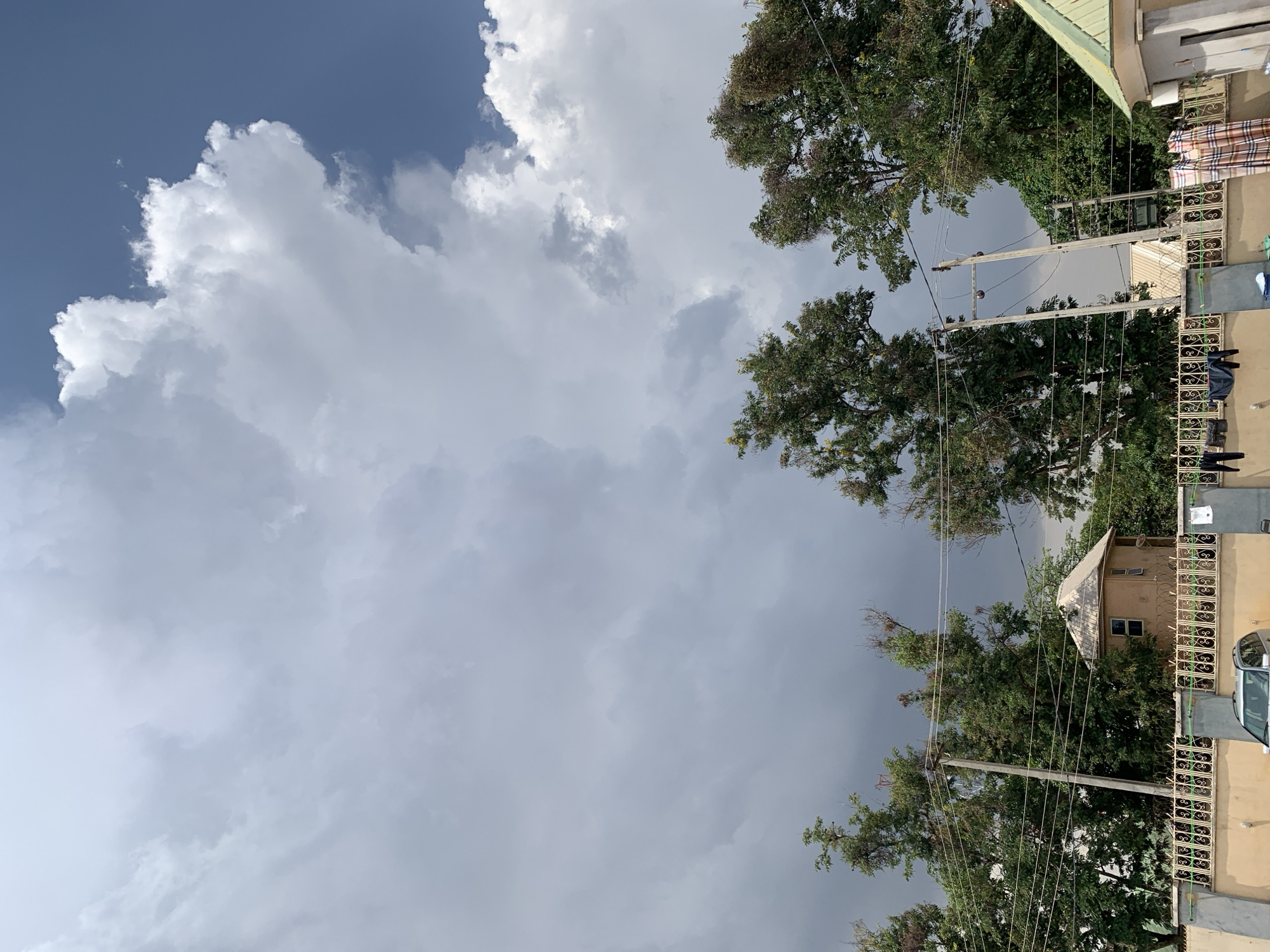 The image shows a view of the sky in Kabong, Nigeria. Cumulus clouds are visible, with some clear blue sky in the upper left corner. The right side of the image contains trees, electrical poles with wires, and rooftops of houses, indicating a residential area. Some items of clothing are hanging on a line. A silver car is visible on the right corner of the image.