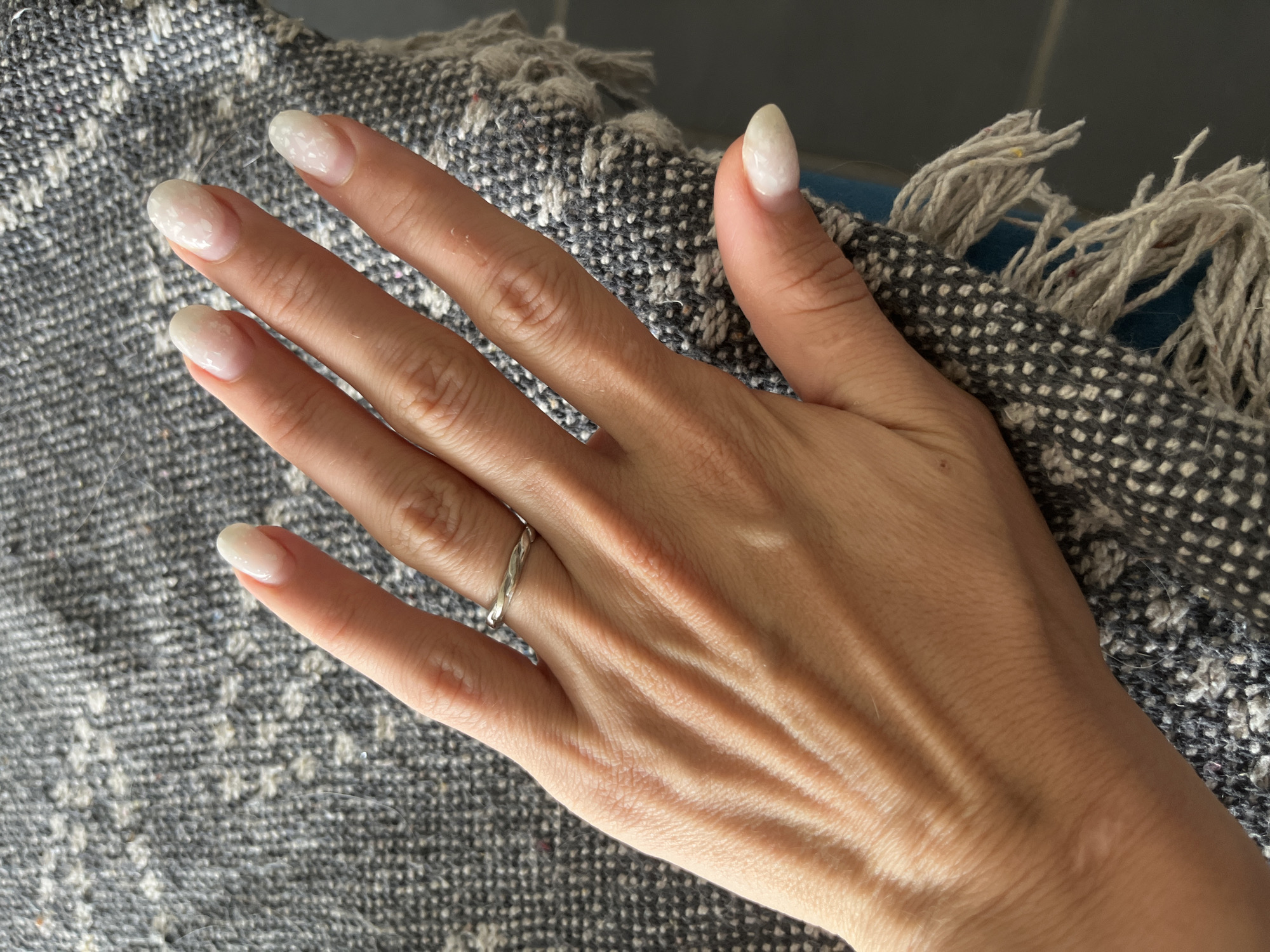 A woman's left hand, with manicured nails and a thin silver ring on her ring finger, rests on a grey, textured fabric with white speckles and frayed edges. The hand is positioned with fingers slightly curled, resting on the fabric which is draped over an unseen surface. The background is out of focus, appearing to be a dark color. The image does not provide any information about a location in Gertwiller, France.