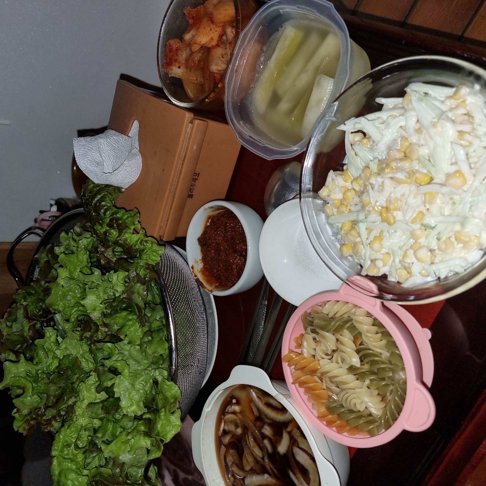 A close-up, eye-level shot captures a spread of Korean side dishes and pasta arranged on a dark wooden table. The primary focus is on a large metal colander overflowing with vibrant green and burgundy lettuce leaves, positioned on the left side of the frame. To the right of the lettuce is a small, light brown rectangular box with Korean text printed on it. Adjacent to the box, a white paper napkin is folded and resting on its edge.
Below the lettuce, a white oval dish contains a dark, savory broth with sliced onions. Next to this dish is a small white bowl filled with a reddish-brown paste, likely a spicy condiment. In the foreground, a pink plastic bowl holds tri-colored fusilli pasta. To the right of the pasta, a clear glass bowl contains a creamy coleslaw mixed with corn kernels. Further to the right, another clear plastic container holds pickled radishes. A small dish of kimchi is visible in the upper right corner. Chopsticks and a spoon are also present on the table. The lighting is dim, suggesting an indoor setting, likely a mealtime. The scene is set in Dunnae-myeon, South Korea.