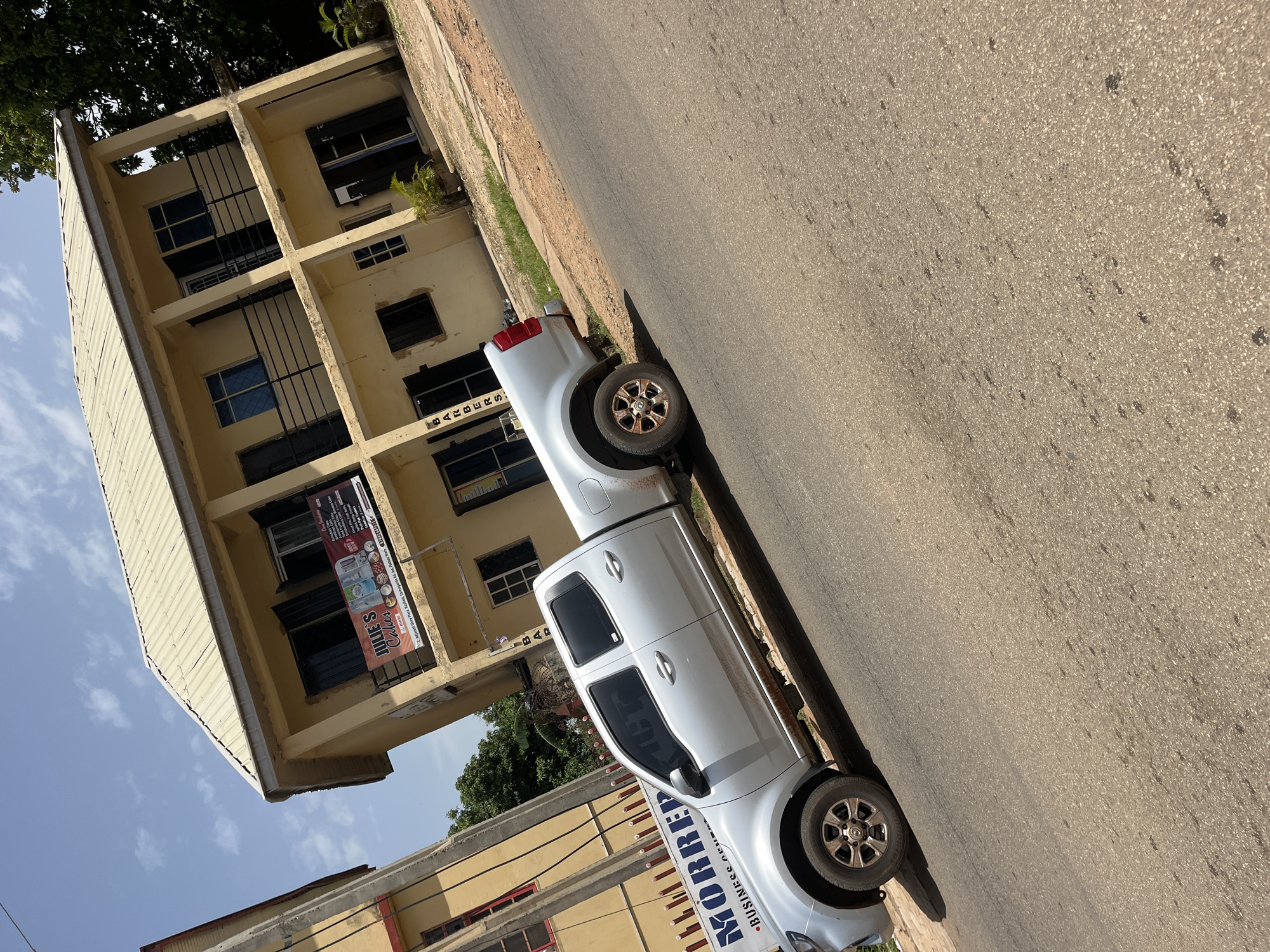 A light silver pickup truck is parked on the side of a paved road in front of a two-story yellow building. The building has several windows and balconies with black railings. A sign on the building reads "JULIE'S CAKES". Another sign partially visible on a fence next to the building says "MORRED BUSINESS AGEN". The scene is outdoors during the day under a clear blue sky with scattered clouds. The ground is asphalt with some dirt and grass on the side. The image appears to be taken in Gura Top, Nigeria.