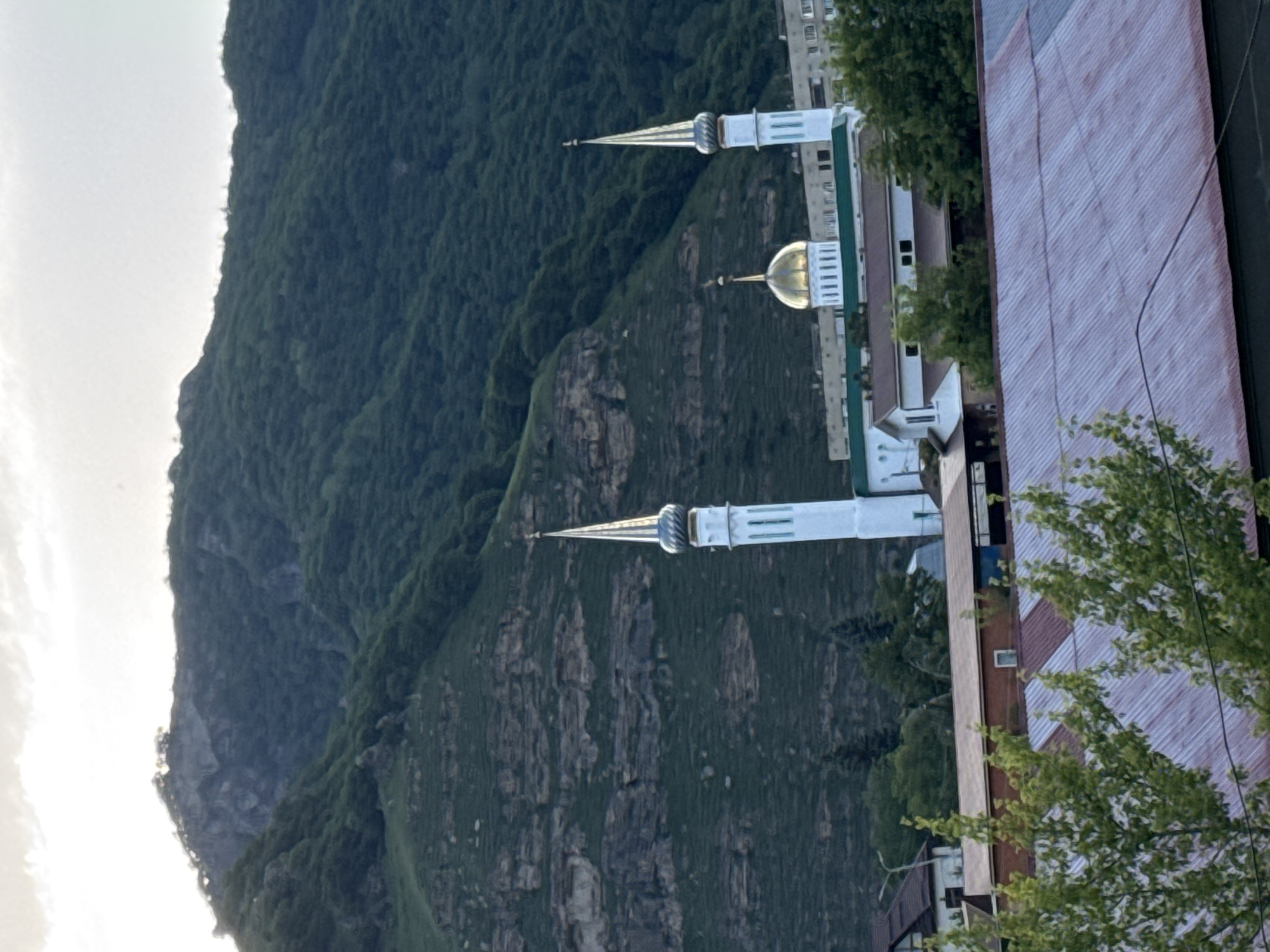 The image shows a view of the city of Karachayevsk, Russia, with a prominent mountain in the background. In the mid-ground, a mosque with a golden dome and two white minarets is visible, along with other buildings and trees. The foreground is dominated by the corrugated metal roof of a building. The sky is overcast.