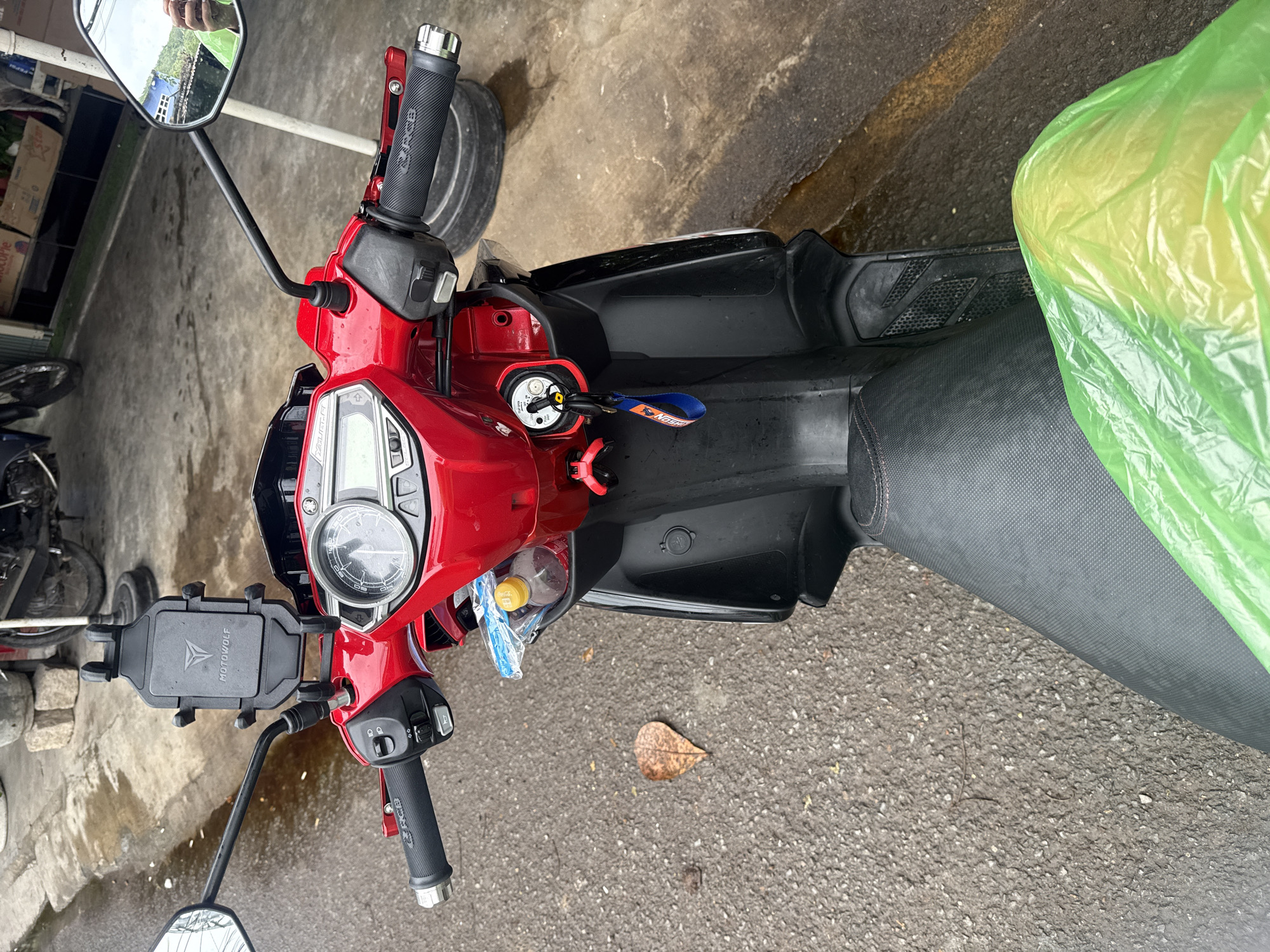 The image captures a close-up, overhead perspective of the handlebars and front section of a red motorcycle parked on a wet asphalt street in Nha Trang, Vietnam. A black "MOTOWOLF" branded phone mount is attached to the handlebars. A motorcycle key with a blue lanyard is in the ignition. To the right of the ignition, a compartment holds a plastic-wrapped bottle of water. A single brown leaf rests on the wet pavement near the front wheel. A partially visible black motorcycle tire and a metallic object are present in the background, suggesting other vehicles or street elements. The overall scene indicates an urban outdoor environment, likely a street or parking area, with signs of recent rain.