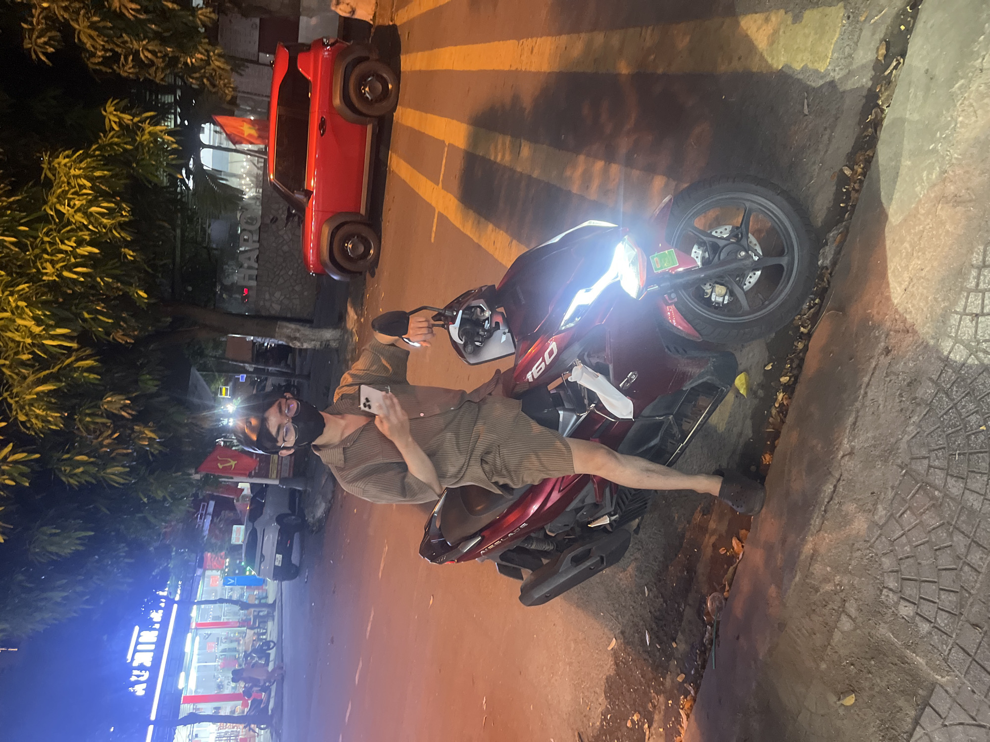 A young Vietnamese person wearing a face mask, glasses, a short-sleeved striped button-up shirt, and striped shorts sits on a dark red scooter at night on a city street in Da Nang, Vietnam. The person holds a smartphone in their right hand and is looking at it. The scooter's headlight is on, illuminating the street. In the background, there are trees, a red SUV parked on the street, and buildings with illuminated signs, suggesting a commercial area. Yellow lines painted on the asphalt mark a crosswalk. The ground is paved with interlocking bricks and asphalt, with some scattered leaves and debris.