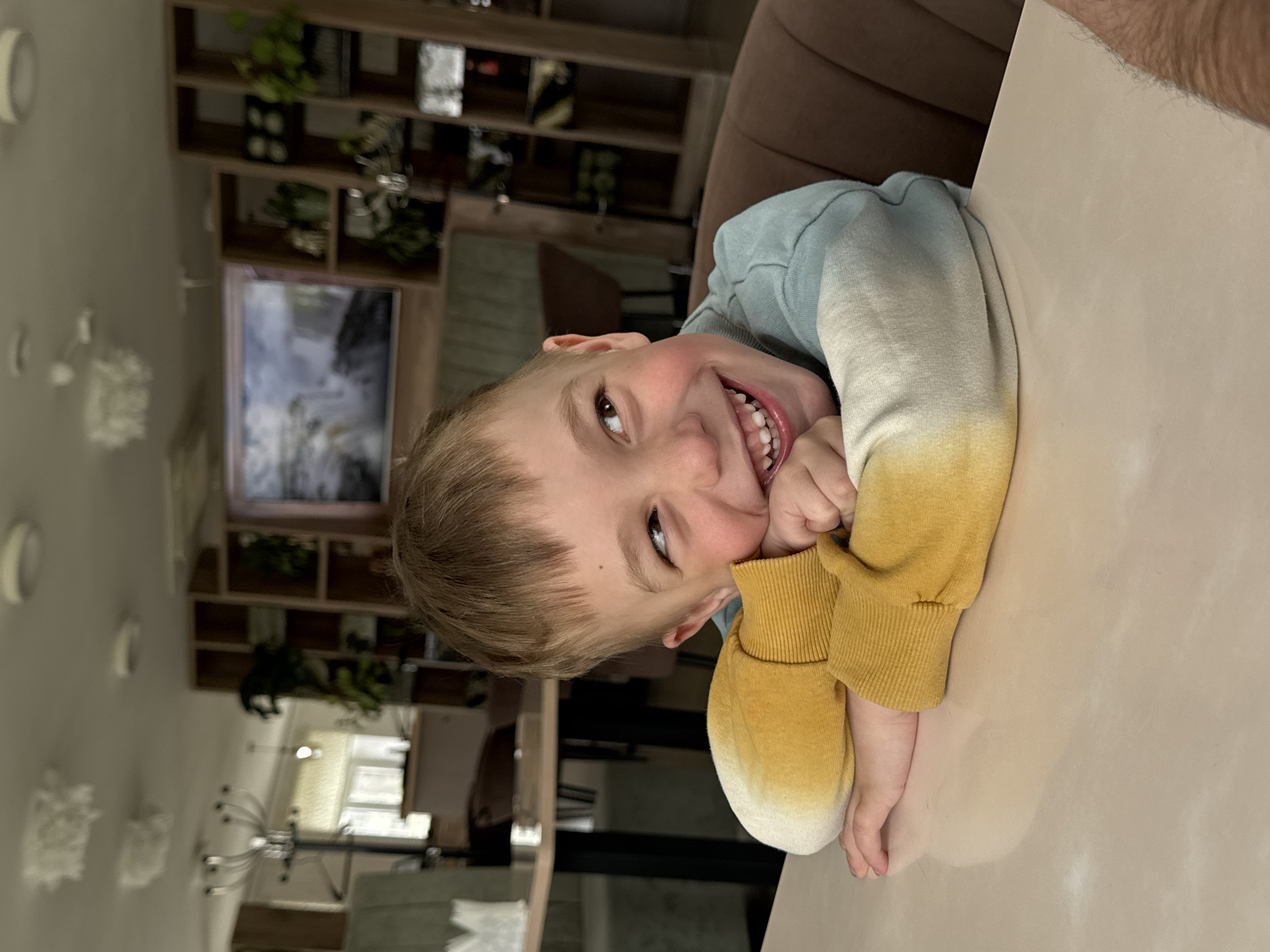 A young boy with light brown hair and fair skin is smiling and resting his cheek on his hands while sitting at a light pink or beige table. He is wearing a blue, white, and yellow tie-dye sweatshirt. The background includes a room with wooden shelves, decorations, a television, and a chandelier-style light fixture. Based on available information about Chaikovsky, Russia, the scene appears to be indoors, possibly in a restaurant or cafe. There is no explicit activity other than the boy posing for the photo.