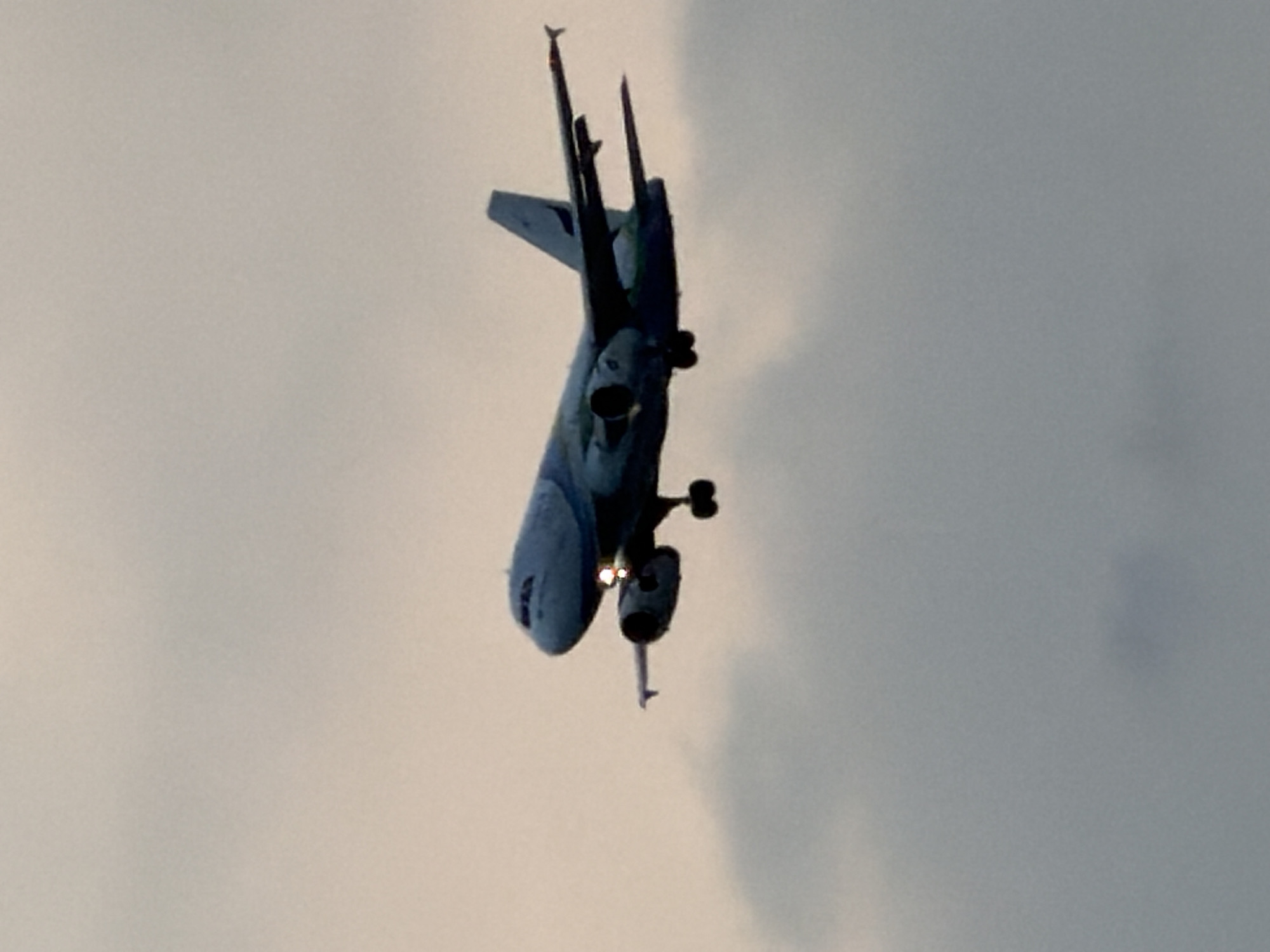 An airplane is captured in mid-flight against a dusky sky, with some clouds visible. The aircraft is a commercial jet with its landing gear extended, suggesting it is either preparing to land or has just taken off. The lighting on the plane indicates it is evening or early morning. The background consists of muted tones of gray and pale yellow, consistent with twilight. There are no discernible landmarks or specific environmental features that would indicate the location within Ko Samui District, Thailand. The image focuses solely on the airplane and the sky.