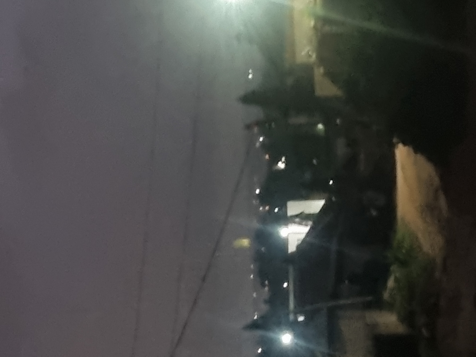 The image depicts a nighttime cityscape, likely in Jos, Nigeria. In the foreground, a dark, blurred foreground suggests a vantage point from a hill or elevated position, looking down upon a settlement. Several structures, possibly houses and buildings, are visible, some with illuminated windows or exterior lights. A dirt road or path winds through the scene. In the middle ground, trees are silhouetted against the night sky, and distant city lights twinkle, indicating a populated area. A prominent, tall, glowing structure, possibly a tower or illuminated building, stands out in the far distance, casting a yellowish light. The sky is overcast and dark, with subtle hints of ambient light. The overall impression is of a quiet, dimly lit urban or semi-urban environment at night.