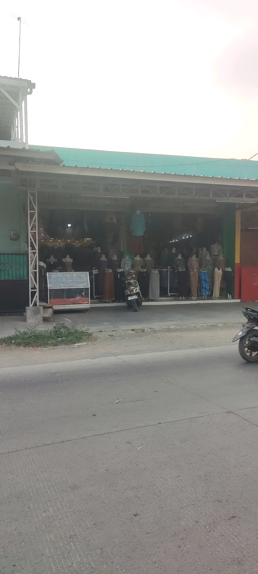 The image depicts a street-level view of a clothing store in Gebang, Indonesia. The store is open-fronted, revealing mannequins dressed in various garments, likely traditional Indonesian clothing such as sarongs and dresses. A motorcycle is parked directly in front of the store. The building has a green corrugated roof and is situated beside a road with a concrete surface. In the background, a white sky suggests it is daytime, possibly late afternoon due to the hazy lighting. The overall scene is a typical street view of a commercial establishment in a small town or city.
