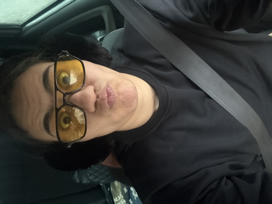 A person with fair skin and dark hair, wearing black earmuffs and yellow-tinted glasses, puckers their lips for a selfie. They are positioned in a car, with a gray seatbelt diagonally across their black crewneck shirt. A quilted patterned item can be seen in the background, likely a car seat or interior detail. The lighting suggests an overcast day.