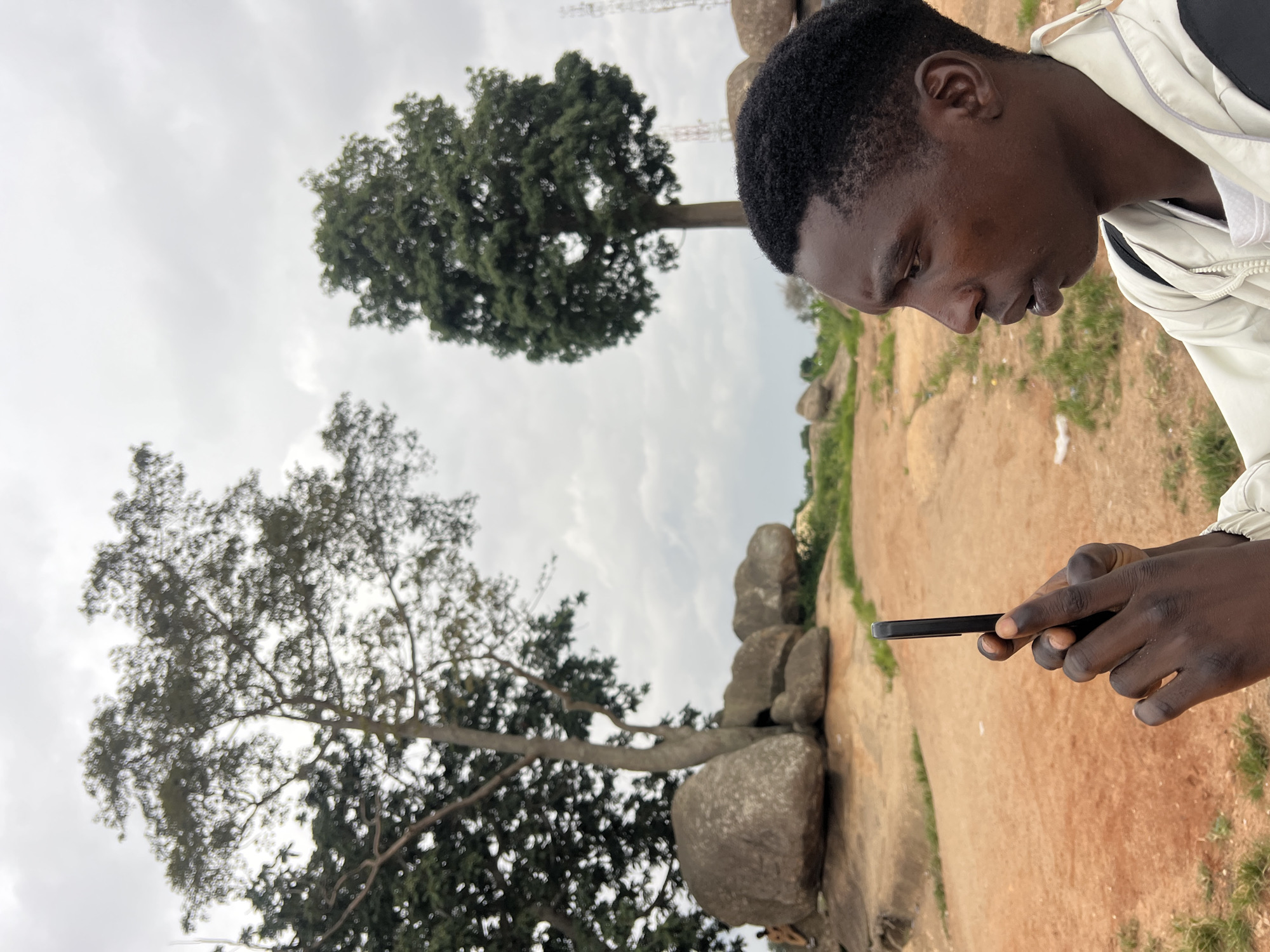 A young Black male in his late teens or early twenties, with dark skin and short, dark, curly hair, is captured in profile, looking to the left. He is holding a black smartphone in his right hand, appearing to take a photograph or video. He is wearing a light-colored jacket over a white t-shirt.
The background consists of a natural outdoor environment. Two large trees are prominent. The one on the left is closer and appears older with a thicker trunk and more extensive branches, partially obscured by smaller, darker green foliage. The tree on the right is taller and more slender with a dense, round canopy of bright green leaves.
Several large, weathered boulders are scattered across the landscape, some resting on a reddish-brown, sandy earth. The terrain is uneven, with patches of dry grass and scrub. The sky is overcast with diffuse, gray clouds. In the far distance, a few communication towers are visible, suggesting the presence of a settlement. The overall impression is a somewhat rugged, undeveloped area.
Based on the characteristics of the landscape, including the unique rock formations and vegetation, this scene is consistent with areas around the city of Jos, Nigeria, known for its rocky terrain and highlands.