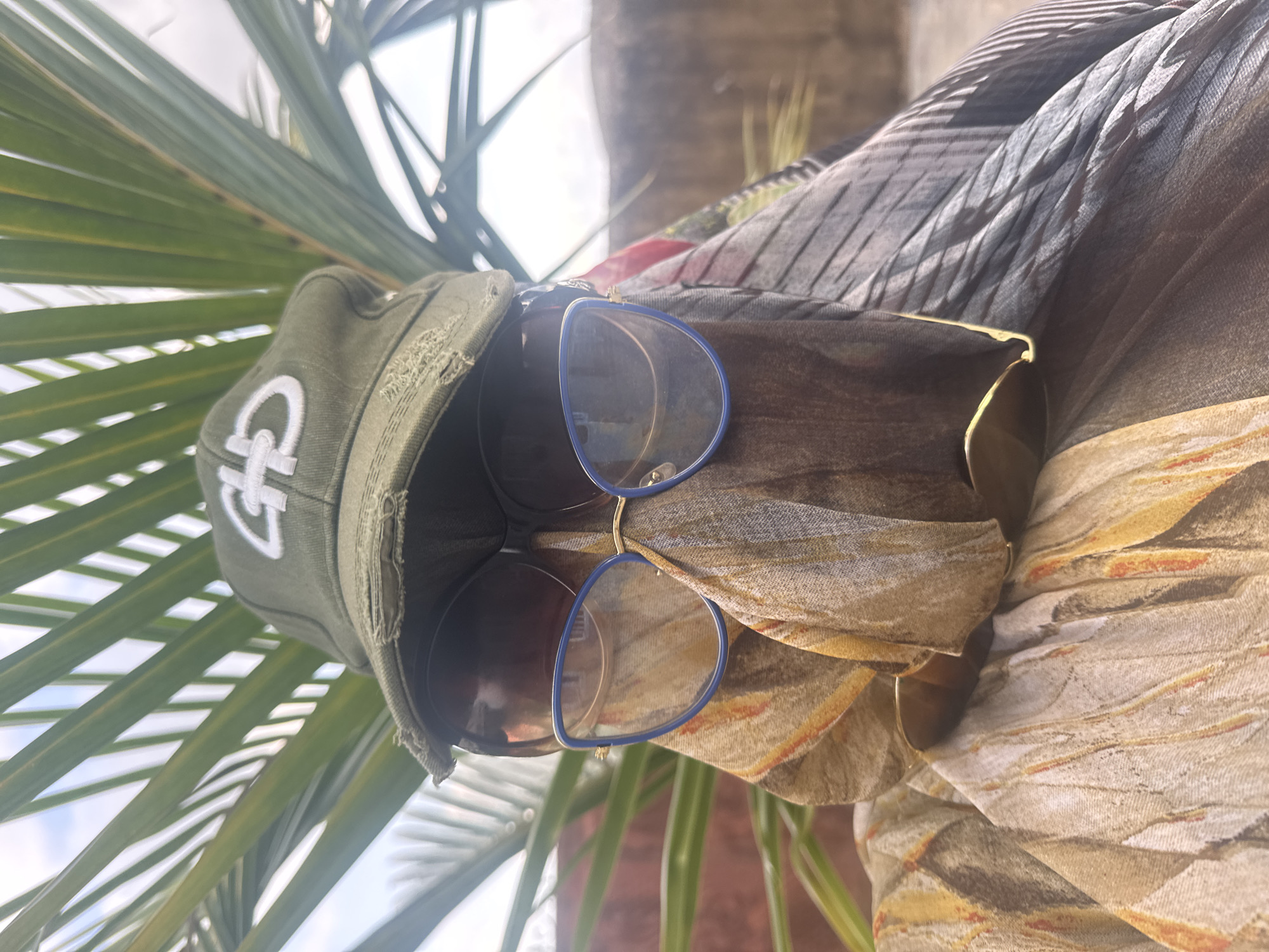 The image is a close-up, eye-level shot of a person wearing a green cap with a white logo, sunglasses with blue frames, and a patterned scarf covering their face and neck. The person is partially obscured by large, green palm fronds that are in the foreground and out of focus. The background consists of a blurred reddish-brown wall and a bright, hazy sky. The scene appears to be outdoors, possibly in a courtyard or garden. There is no specific indication of the location being in Jawu, Nigeria.