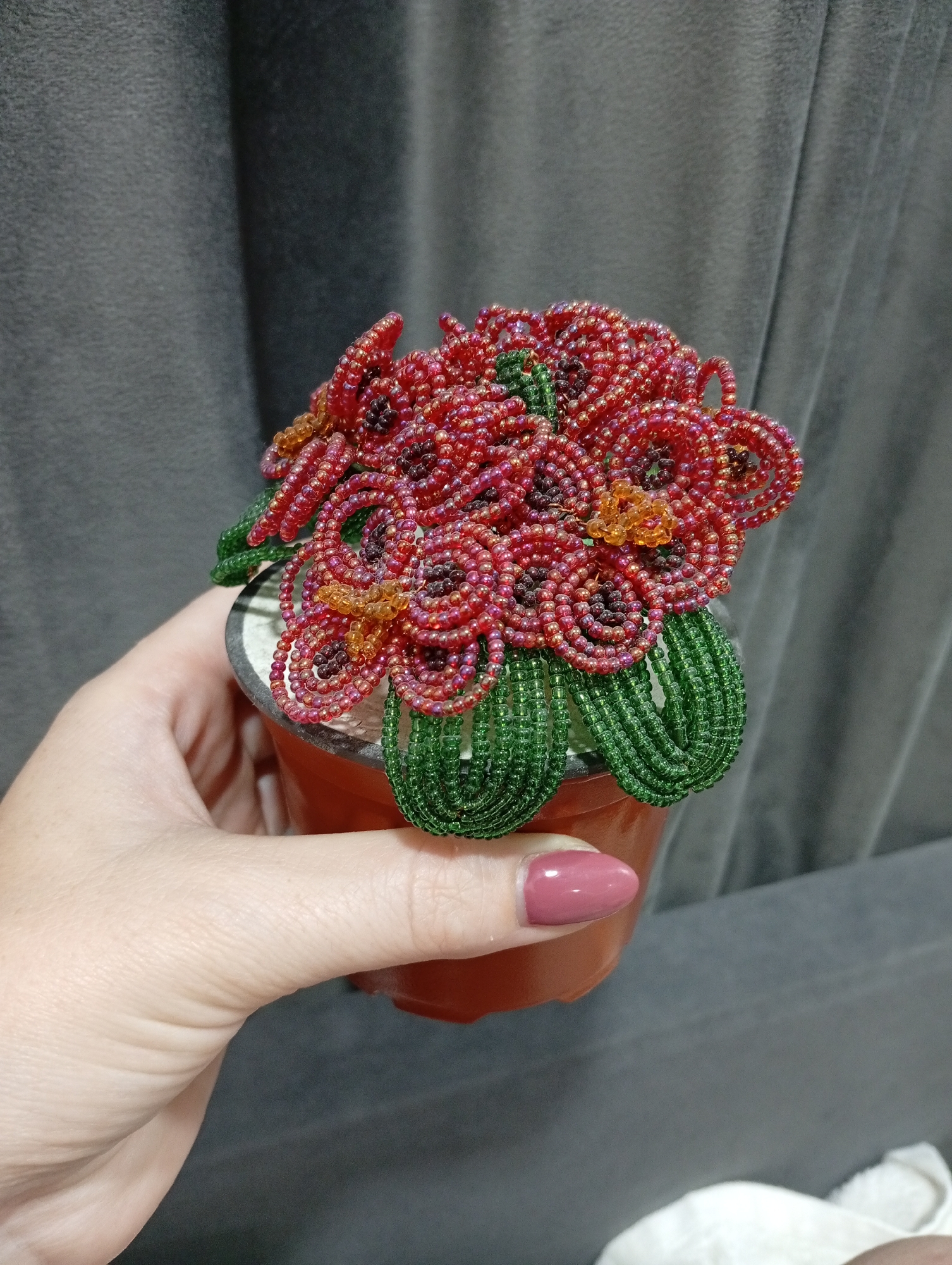 A close-up, eye-level shot shows a hand holding a small, orange pot containing a beaded craft plant with red flowers and green leaves. The background is a grey, textured curtain. The hand has light skin and pale pink nail polish. The plant is made of tiny red, orange, and dark purple beads for the flowers, and green beads for the leaves. It is in the city of Pănășești, Moldova.