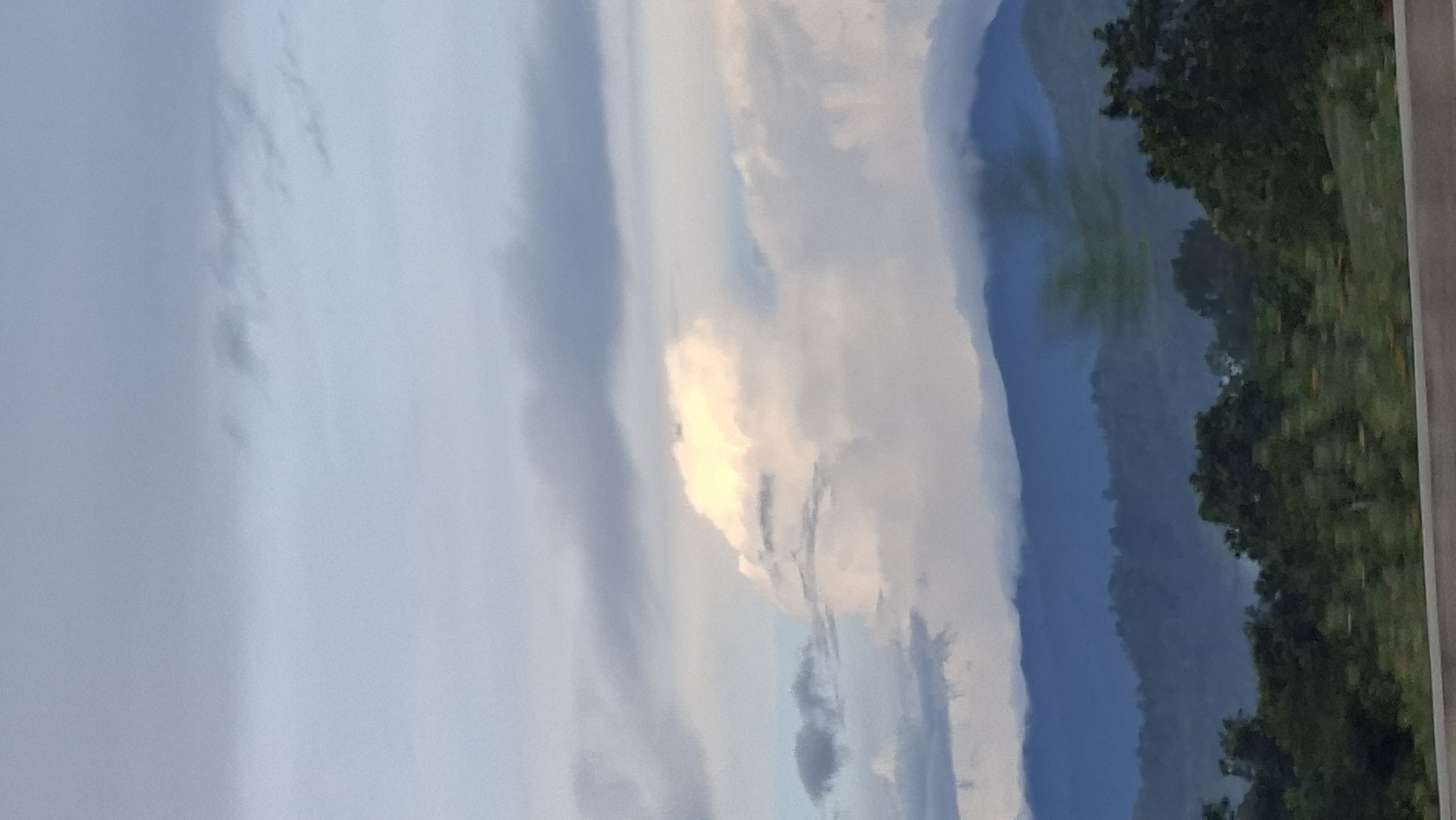 The image shows a hazy, distant view of mountains and trees, likely captured from a moving vehicle. The sky is overcast with various shades of gray and white clouds, with a bright patch of light suggesting the sun's presence behind the clouds. The mountains in the background are a deep blue, with a lighter mist or cloud cover obscuring their peaks and lower slopes. In the foreground, there are dark green trees, some of which appear blurred due to motion. A gray horizontal line runs across the bottom of the image, possibly representing the edge of a road or barrier. The overall atmosphere is muted and slightly diffused. The scene is consistent with a view from a highway or elevated road in the region of Lamtamot, Indonesia, where such mountainous terrain and tropical vegetation are common. There are no people or specific identifiable structures visible.