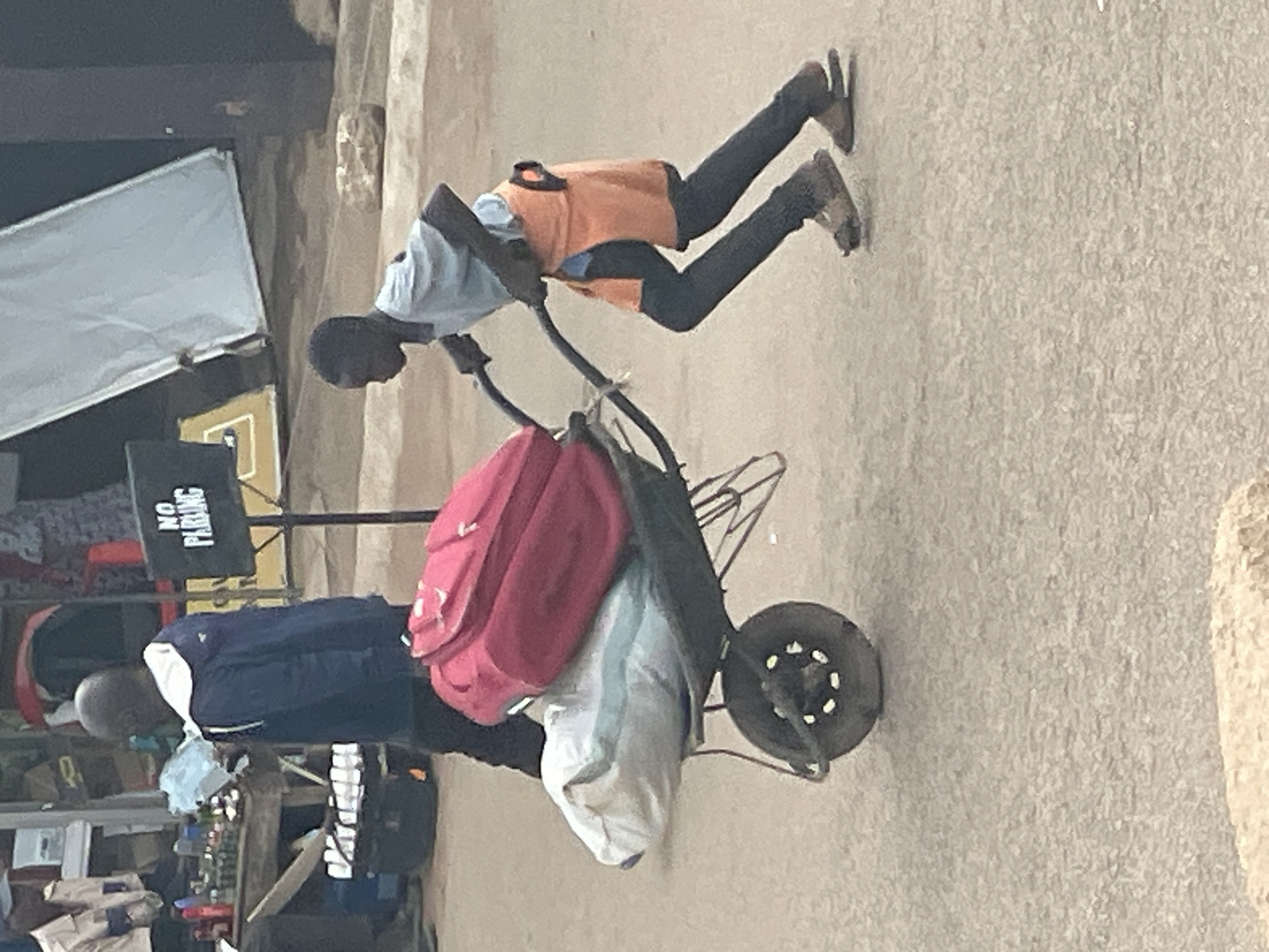 A young boy pushes a wheelbarrow loaded with two large bags, one red and one light blue, on a paved street. He is wearing a light blue t-shirt, an orange skirt over dark leggings, and sandals. The wheelbarrow has a single wheel and two handles. In the background, a man in a blue jacket walks away from the camera. There is a "NO PARKING" sign and a market stall with various items visible. The setting appears to be an outdoor urban environment.