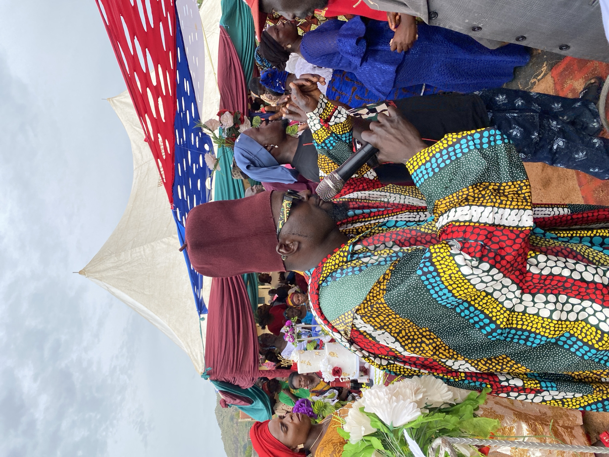 The image depicts an outdoor event, possibly a wedding or celebration, in Kuru Dungus, Nigeria. A man in a colorful patterned shirt and maroon fez is speaking into a microphone. The background reveals a crowd of people under a canopy adorned with red, white, and blue fabric, as well as maroon and green drapes. There's a tiered white cake visible in the background. The sky appears overcast. People are gathered, seemingly listening to the speaker. In the foreground are white flowers.