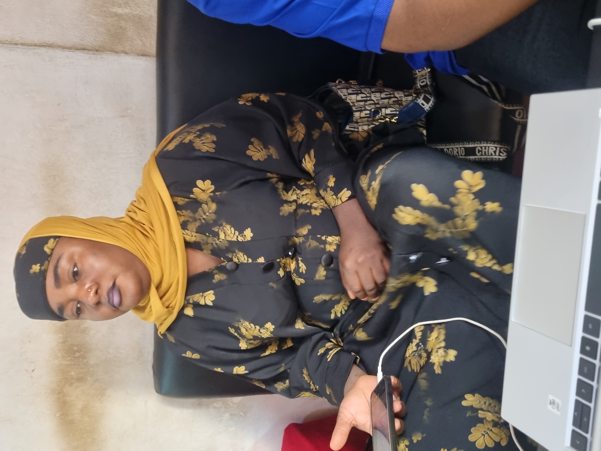 The image depicts a woman seated on a black leather couch, wearing a black and gold patterned dress and a yellow hijab. She has dark skin, is wearing purple lipstick and a nose ring. In her left hand, she holds a smartphone. A white charging cable extends from the phone. To her right, a portion of a person wearing a blue shirt is visible. In the foreground, a silver laptop is positioned on a surface. A handbag with a "Christian Dior" pattern is visible on the couch to the woman's right. The background consists of a textured, light-colored wall. The scene appears to be an indoor setting, possibly a lounge or living area, located in Jos, Nigeria.