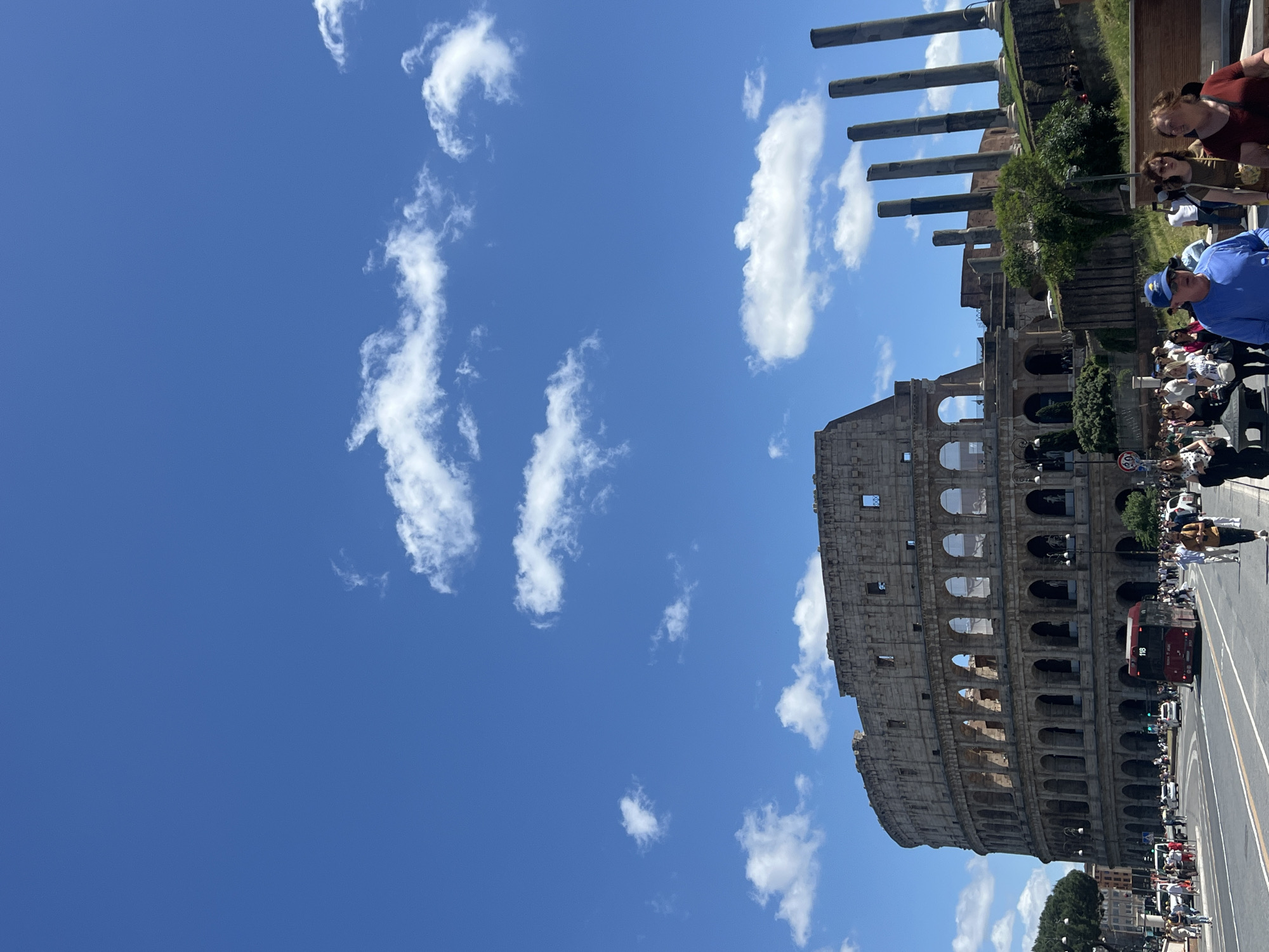 The image shows a view of the Colosseum in Rome, Italy on a sunny day. The sky is bright blue with scattered white clouds. The Colosseum is made of stone and has multiple tiers of arches. People are walking and standing near the Colosseum, with a double-decker bus and other vehicles on the road in front. Ruins of other structures are visible adjacent to the Colosseum, including several tall columns.