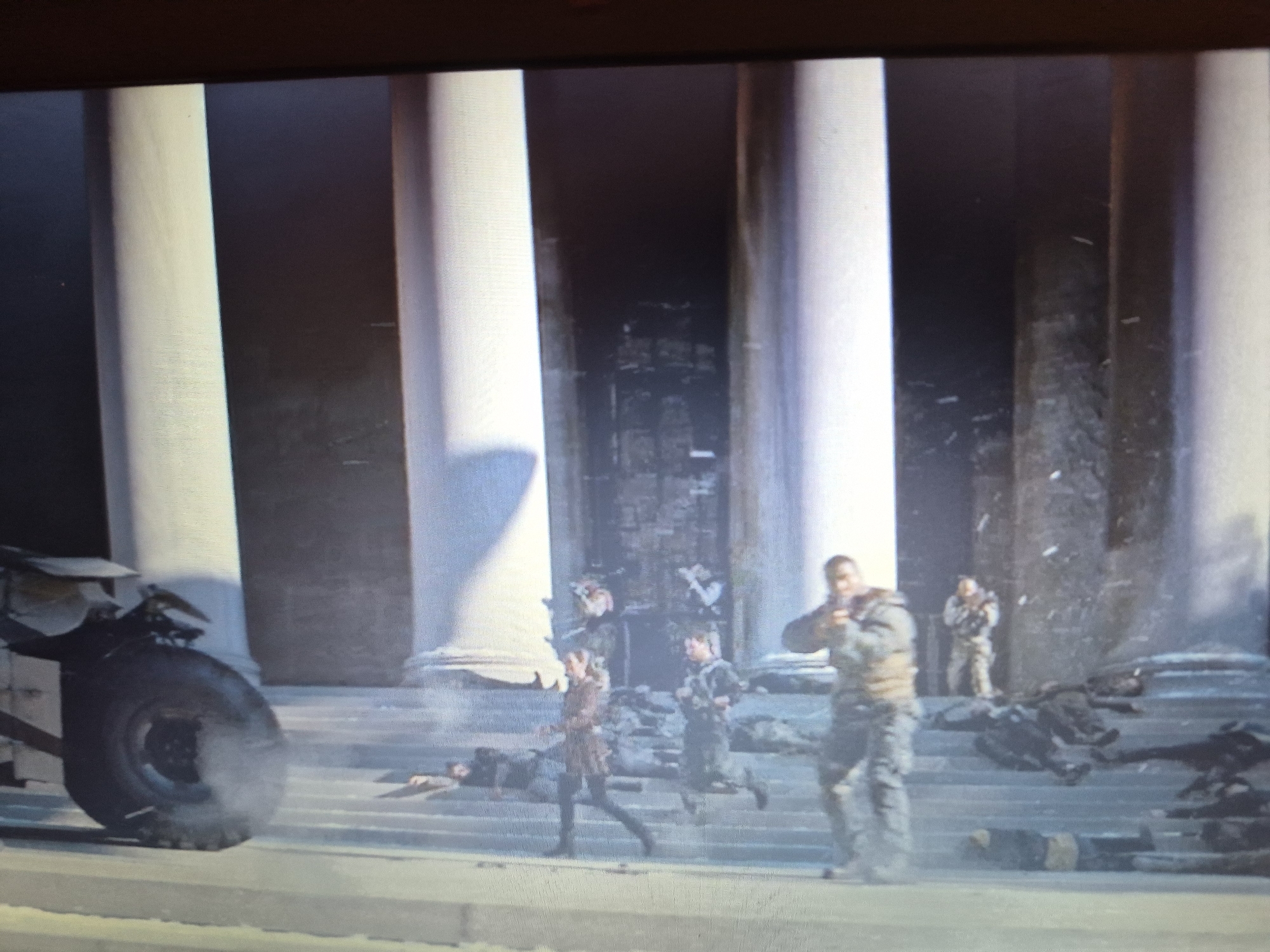 The image depicts a chaotic scene in front of a grand, classical-style building with large columns. Several figures in military attire are visible. In the foreground, a large vehicle wheel, possibly from a tank or armored car, is prominently featured and appears to be emitting smoke. The ground is littered with numerous fallen bodies, suggesting a recent conflict or battle.
In the mid-ground, several individuals in military uniforms are seen moving. One person in the foreground is standing and aiming what appears to be a weapon. Another figure in military uniform is running up steps. In the background, more individuals in military attire are present, and the entrance to the building appears damaged.
There is no discernible information within the image to suggest the scene is located in the city of Sandwell, United Kingdom. The architectural style and the presence of military personnel do not provide any specific geographical markers for Sandwell.