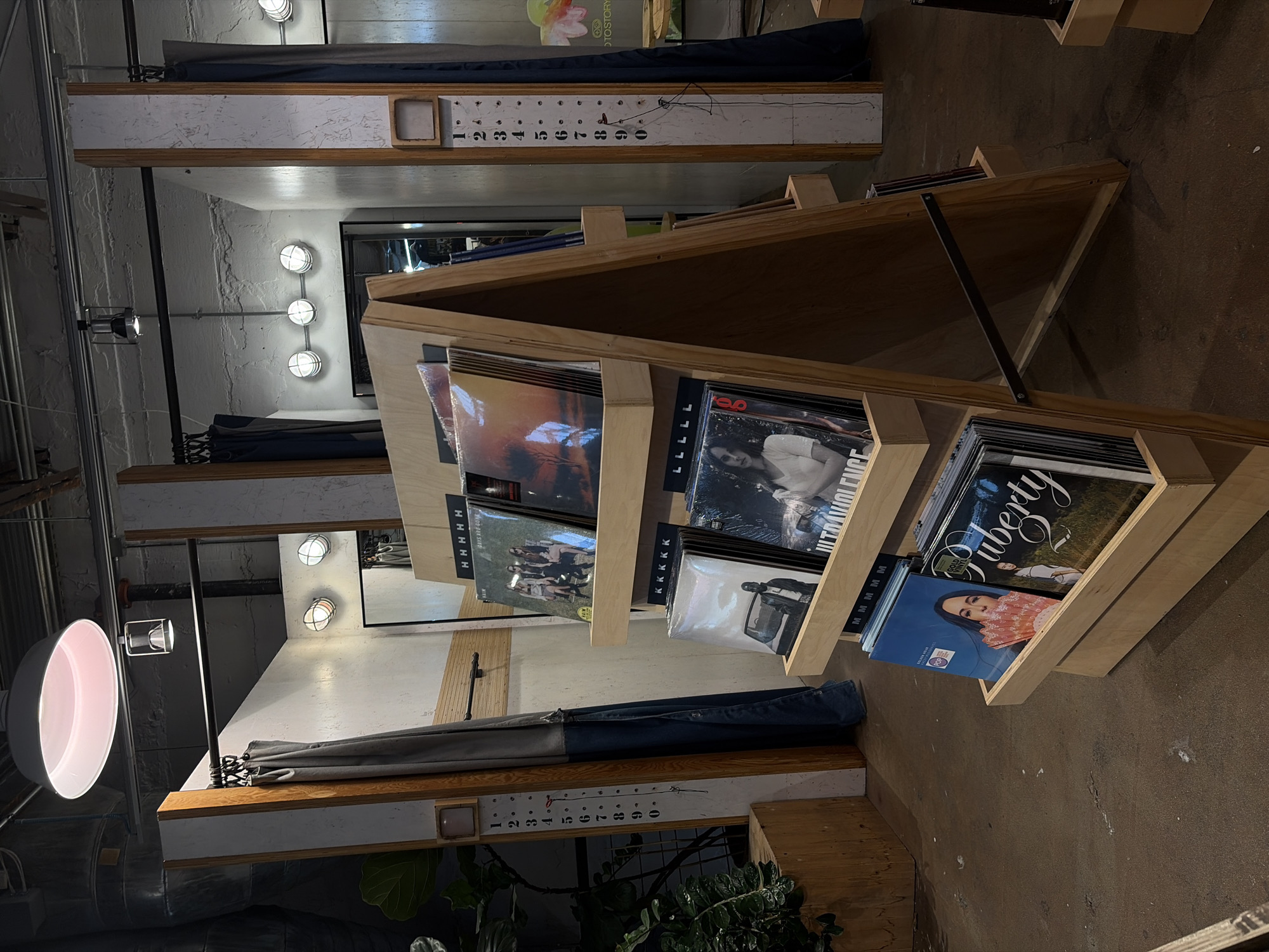 This indoor shot displays a record store environment, likely located in Austin, Texas, United States. The main focus is a wooden record display rack that is angled, showing album covers in each section. Some of the album covers are visible, containing words like "ULTRAVIOLENCE" and "puberty."
The display rack sits on a concrete floor. Behind the rack, the architectural details of the building are visible, showing white walls and wooden support beams. The number of the records displayed are marked as "H", "K", and "M". A gray material appears to be hanging from the beam.
The building has industrial elements. The lighting is provided by multiple round fixtures affixed to the walls and a large round lamp to the left, adding to the ambience. The color scheme is neutral with wood tones, white, and gray. A plant is shown on the floor as well.