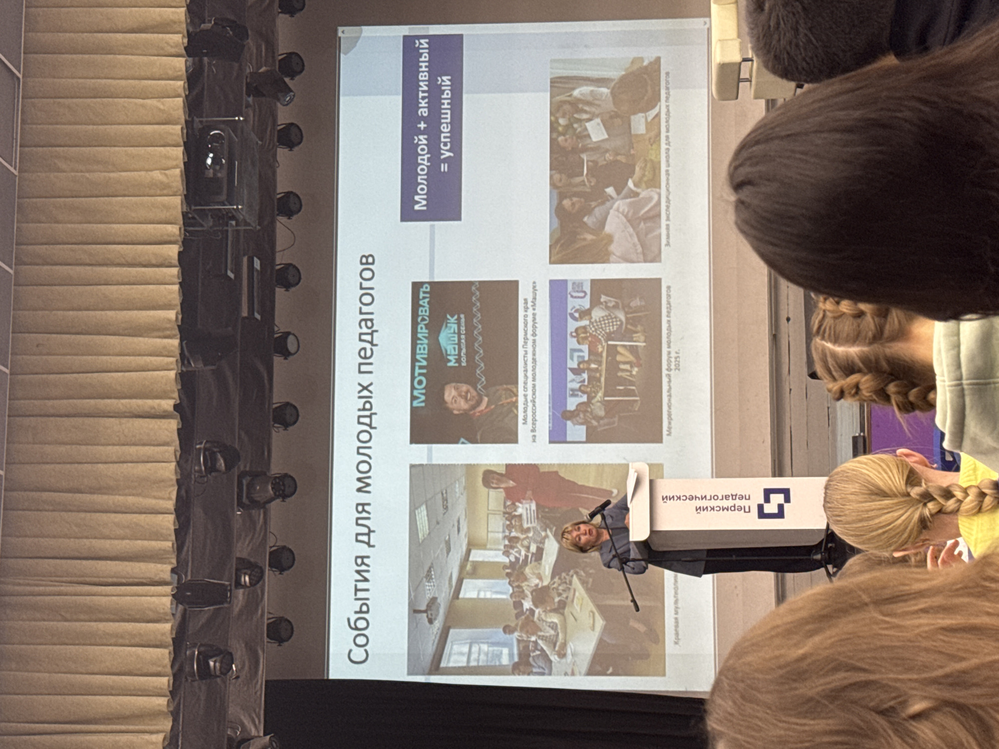 The image captures an indoor educational event, likely a conference or presentation, taking place in Perm, Russia. A speaker stands behind a podium, which is branded with "Пермский педагогический" (Permsky pedagogical), indicating a connection to a Perm-based pedagogical institution or event. The speaker is addressing an audience, whose heads and shoulders are visible in the foreground, suggesting they are seated and listening to the presentation.
A large projection screen dominates the background, displaying a slide titled "События для молодых педагогов" (Events for young educators). The slide is divided into sections showcasing various programs and initiatives. One prominent section features the text "МОТИВИРОВАТЬ" (Motivate) alongside the logo "Машук БОЛЬШАЯ СЕМЬЯ" (Mashuk BIG FAMILY), with details about "Молодые специалисты Пермского края на Всероссийском молодежном форуме «Машук»" (Young specialists of Perm region at the All-Russian youth forum "Mashuk"). Another section emphasizes the equation "Молодой + активный = успешный" (Young + active = successful).
Three illustrative images are present on the slide:
1. An image on the far left shows a speaker addressing a room filled with young individuals at tables, potentially representing a "Краевая мультиолим" (Regional multi-olymp).
2. Below the "МОТИВИРОВАТЬ" section, an image depicts participants engaged in a group activity, identified as the "Межрегиональный форум молодых педагогов 2025 г." (Interregional forum for young educators 2025).
3. On the bottom right, another group image is captioned "Зимняя экспедиционная школа для молодых педагогов" (Winter expeditionary school for young educators).
The setting includes a series of stage lights mounted along the left side of the presentation area. The overall scene suggests a professional development or networking event aimed at empowering and engaging young educators.