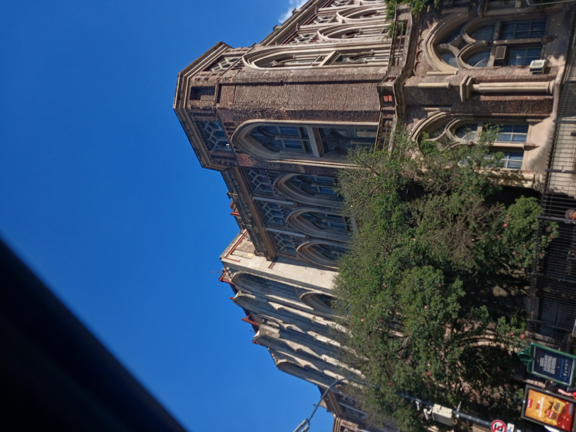 The image shows a building, presumably the Church of the Holy Cross, located in Buenos Aires, Argentina. It is a multi-story building with gothic-style architecture, featuring arched windows and intricate stonework. The building is made of brick and stone, with some of the facade covered by green foliage from trees lining the street. A portion of the sky is visible, indicating it is a bright, sunny day. Street furniture, including signs, can be seen near the base of the building, indicating it is an urban environment. The image is rotated.