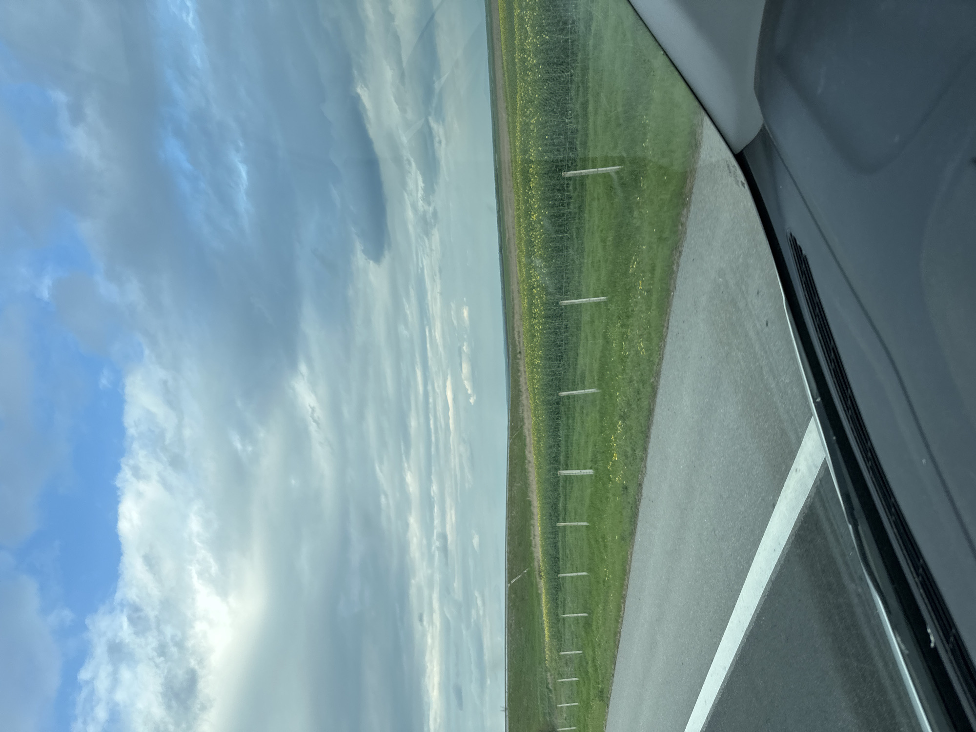 The view from inside a vehicle shows a roadway with a white line marking the edge. To the left of the road is a grassy field with evenly spaced white posts along the perimeter. Beyond the field, the sky is visible with a mix of blue and white clouds. The interior of the car is partially visible on the right side of the image. The location is likely near Загорци, Bulgaria, due to the environment appearing to be typical of that area.