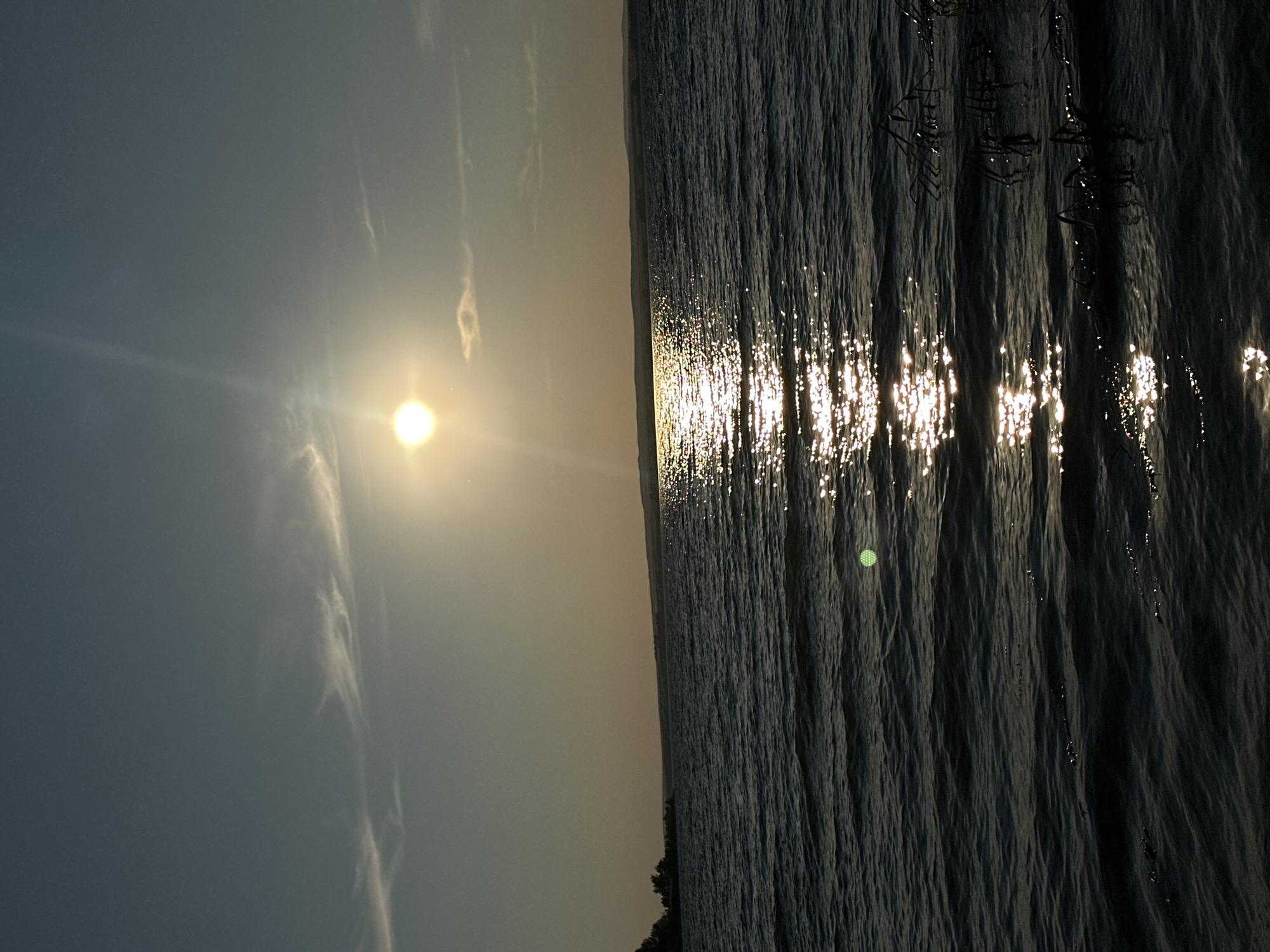 The image depicts a body of water, likely a lake or large river, with the sun low in the sky, casting a strong reflection on the rippling surface. The sky is a gradient of blue and yellow, with thin wisps of clouds. In the distance, a dark, low-lying horizon is visible, possibly indicating land or hills. On the left side of the image, a silhouette of trees can be seen against the sky. The foreground consists of dark, choppy water with some sparse vegetation emerging from it on the right side. The overall impression is that of a tranquil, natural scene, possibly captured during a sunset or sunrise. The location is identified as the city of Energetik, Russia, which is known for its proximity to Lake Baikal.