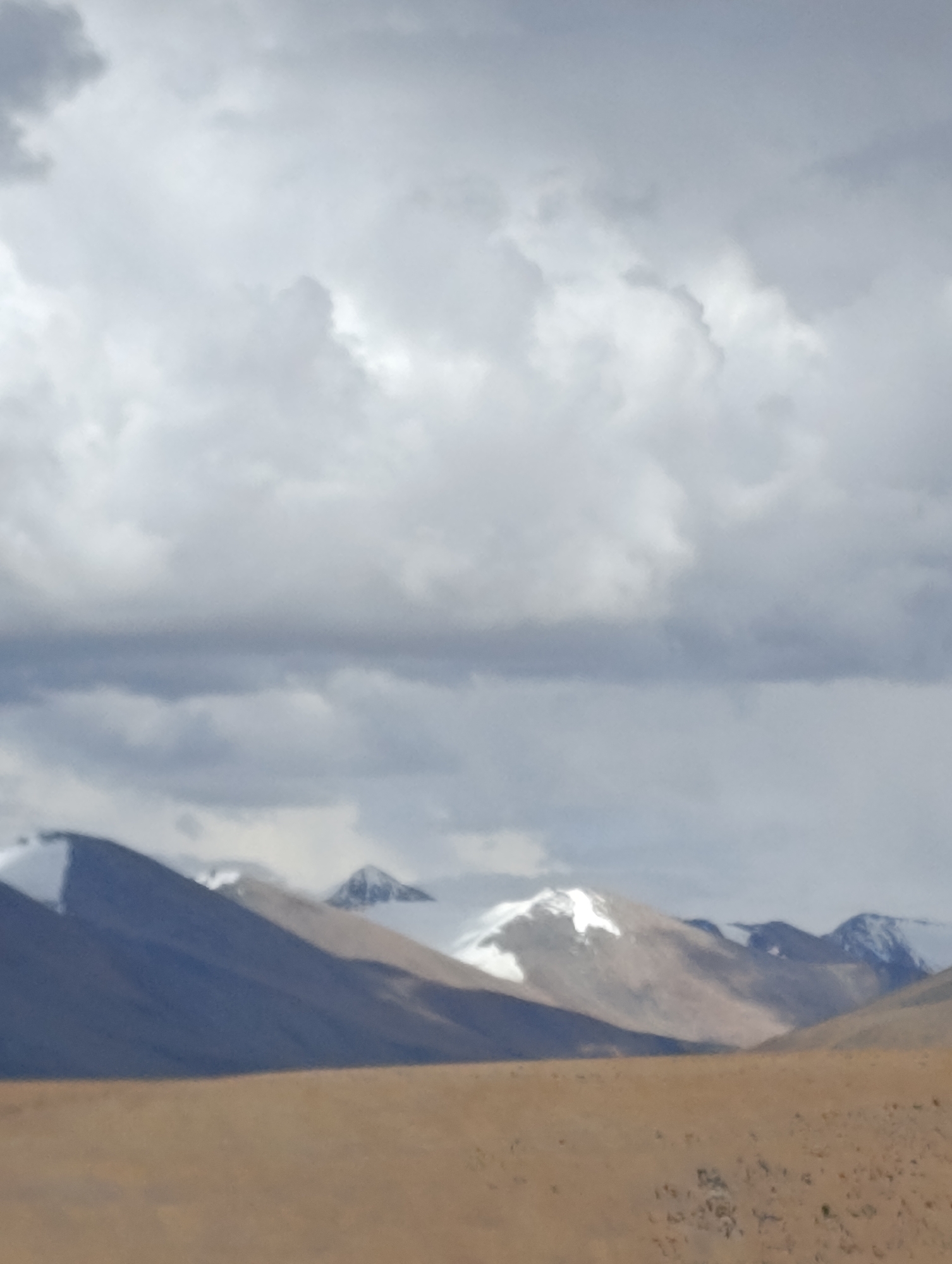 This is a vertical, medium-shot, eye-level photograph of a vast, mountainous landscape under a cloudy sky. The foreground is dominated by a dry, brown, desert-like plain with sparse vegetation. Behind the plain, a range of imposing mountains rises, their peaks dusted with snow. The mountains are in varying shades of brown, grey, and dark blue, with shadows cast across their slopes, indicating the position of the sun. The sky above is filled with a thick layer of grey and white clouds, some of which are dark and ominous, suggesting an overcast day. The overall impression is one of rugged, remote, and untamed wilderness. The location is described as Unknown, India, suggesting a remote or unspecific geographical setting within India. There are no people, animals, or man-made structures visible in the scene.