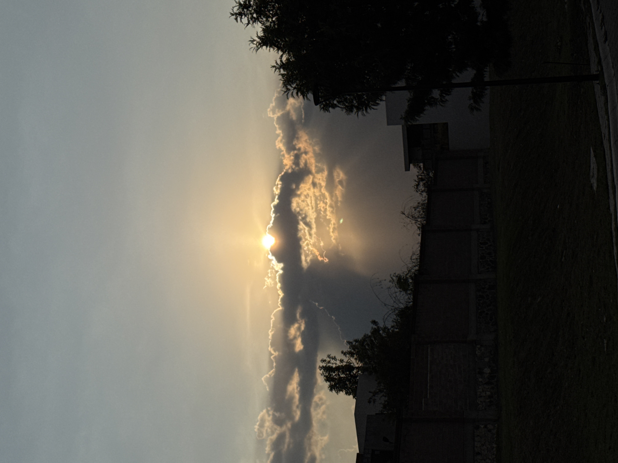 The image depicts a view towards the sky, partially obscured by clouds, with the sun breaking through. The light casts a yellowish glow on the clouds and surrounding sky. Silhouetted trees and parts of buildings are visible along the right side of the frame. A fence can be seen below one of the buildings on the right. The location is Santa Clara Ocoyucan, Mexico.