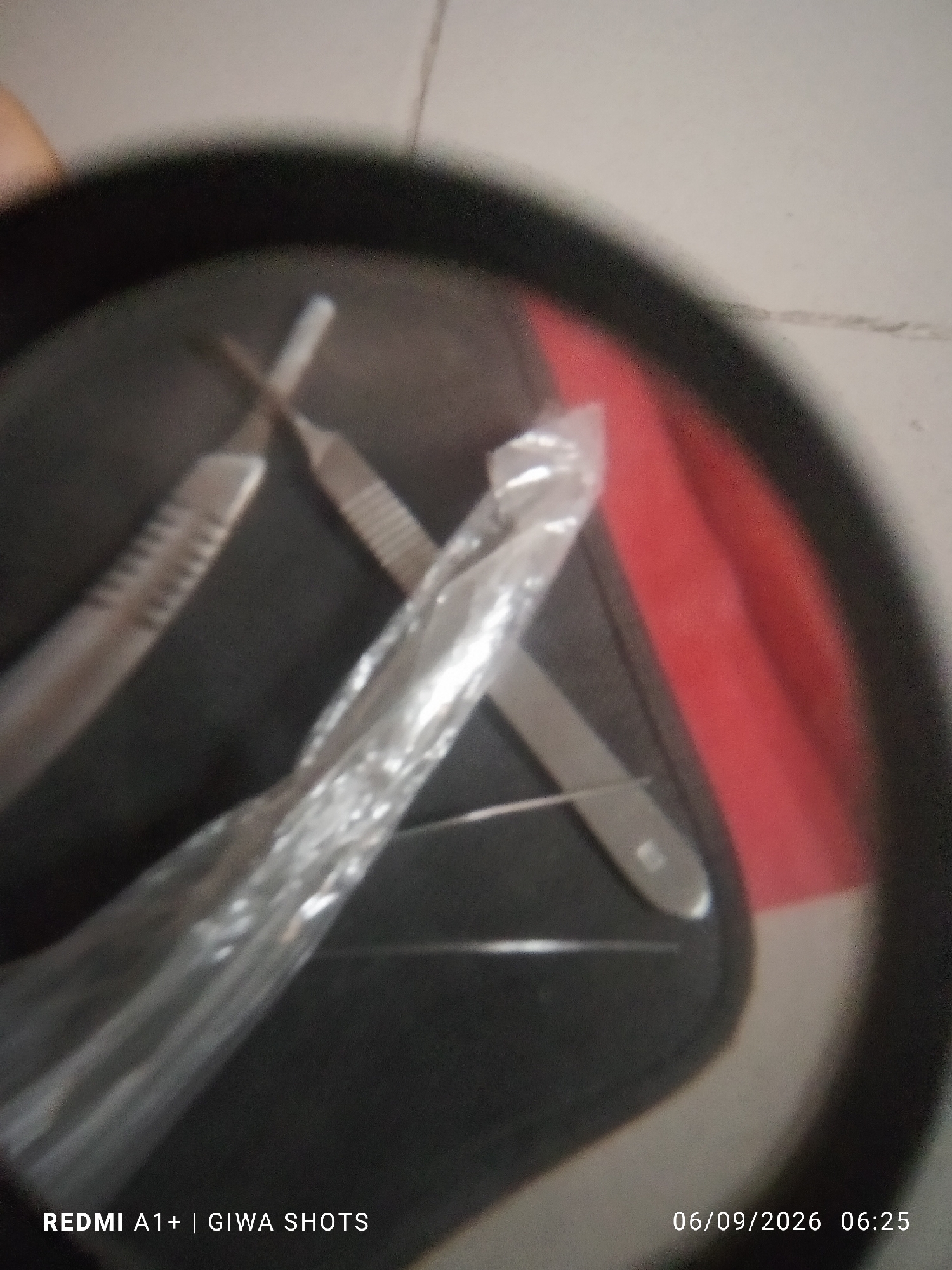The image, taken in an unknown city and location, shows a close-up view of medical instruments, possibly scalpels, arranged on a dark surface with a red accent. Two of the instruments appear to be crossed. One instrument is partially obscured by clear plastic wrapping, which also has some reflective highlights. A fine, thin needle or wire is visible underneath the plastic. The overall image is slightly blurry, suggesting it was taken with a handheld device or in low light conditions. A timestamp in the lower right corner reads "06/09/2026 06:25", and text in the lower left corner indicates "REDMI A1+ | GIWA SHOTS".