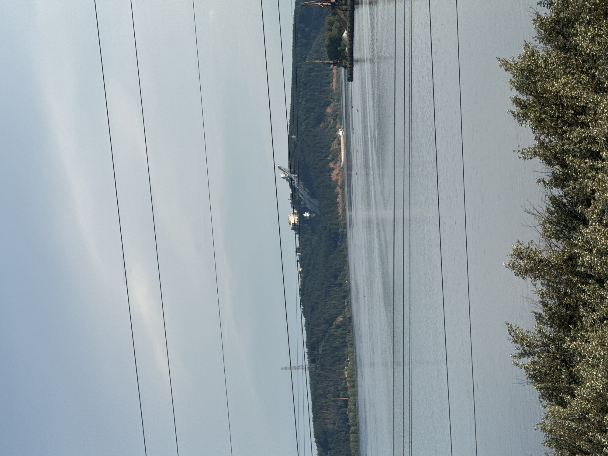 The image shows a wide river with a wooded hill on the opposite bank. A large industrial conveyor belt system is visible on top of the hill, along with some buildings. A barge is moving on the river. In the foreground, there are trees. The sky is overcast. The scene is consistent with a view of the Kama River in Chaikovsky, Russia, near the port and the Kamsky Pulp and Paper Mill.