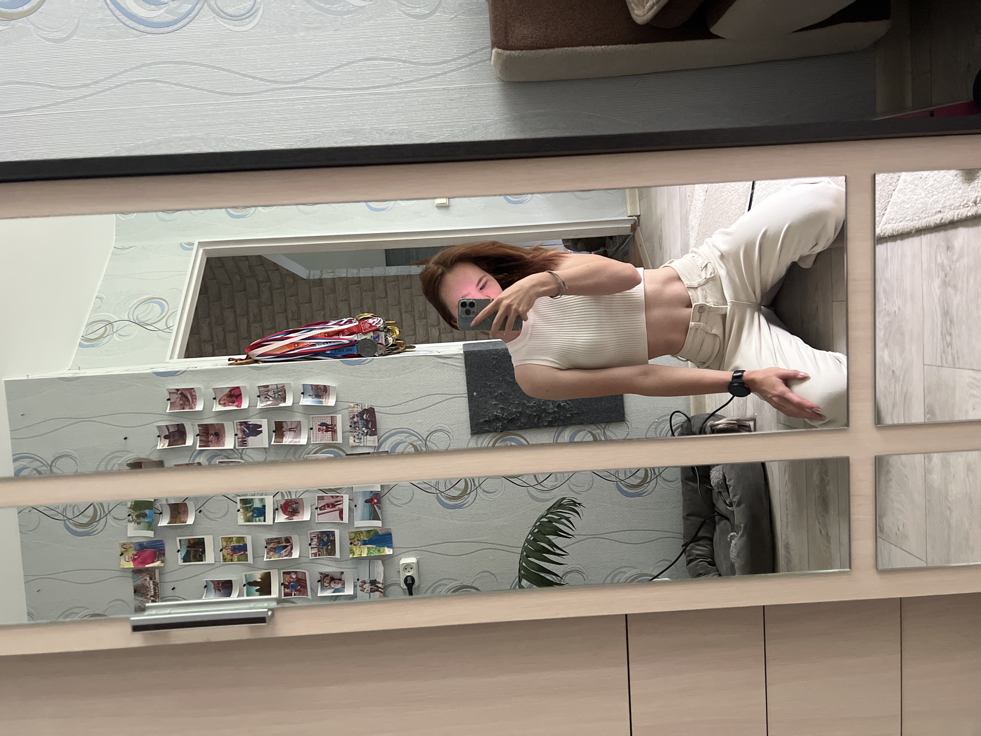A young woman with auburn hair is kneeling on the floor, taking a mirror selfie. She is wearing a white ribbed tank top and loose-fitting cream-colored pants. Her phone is held up to her face, obscuring it partially. Behind her, the mirror reflects a room with patterned wallpaper, framed photos on the wall, a doorway leading to another room, a pet bed with a dog lying in it, and medals hanging on the wall. The room appears to be a bedroom or living area in a residential apartment. There is a wardrobe to the left of the mirror, and part of a bed with a brown headboard is visible to the right. The floor is covered with light-colored laminate flooring and a rug. This scene is likely located in an apartment in Chaikovsky, Russia, based on the prompt.
