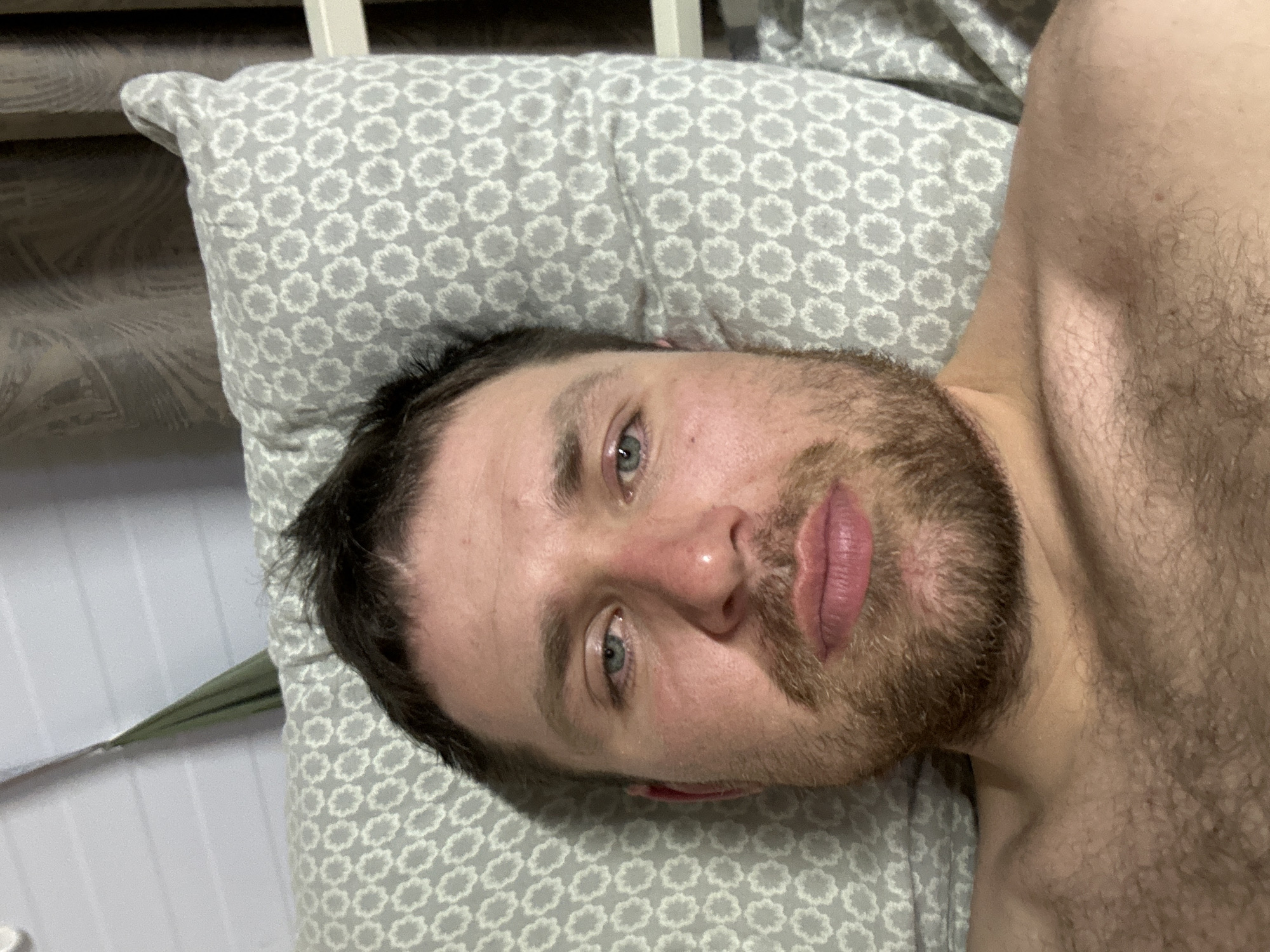 A Caucasian male with a beard and short, dark hair is lying on a pillow with a gray and white floral pattern. He appears to be resting in a supine position. His chest and upper torso are visible, suggesting he is shirtless. The background shows part of a white bed frame. There is no visual evidence to indicate this image was taken in Nizhnekamsk, Russia.