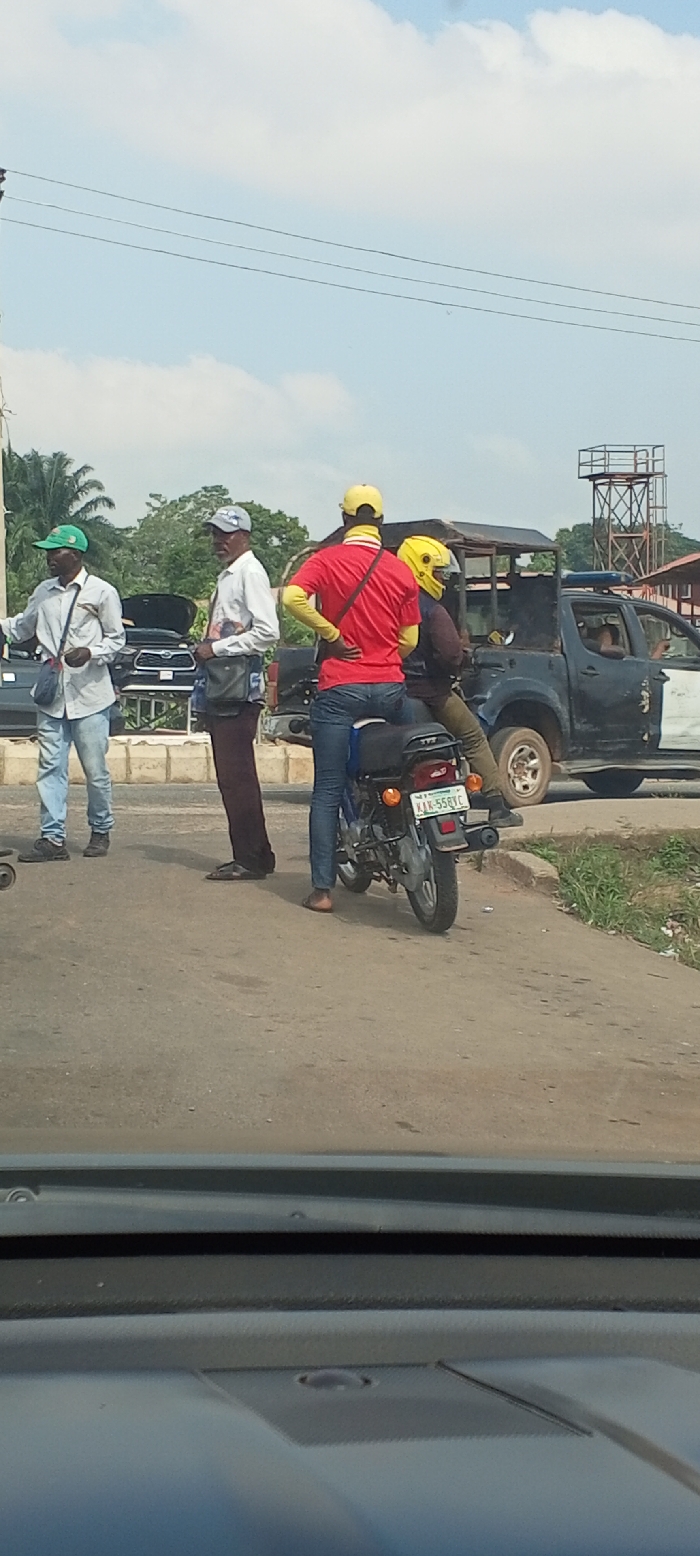 An outdoor scene in Akure, Nigeria, depicts a roadside interaction under a bright, cloudy sky. Four adult males are prominent. On the left, two dark-complexioned men stand by the road's edge. The leftmost wears a green cap, light shirt, and blue jeans, carrying a light blue sling bag. The man beside him wears a grey cap, white graphic shirt, and dark trousers, with a dark sling bag. To their right, a dark-colored TVS motorcycle, with Nigerian license plate KJK-558XC, is stopped. Its driver, seen from behind, wears a yellow cap, a red shirt over a yellow long-sleeved shirt, and blue jeans, with a black sling bag. A passenger, wearing a bright yellow full-face helmet, dark jacket, and dark trousers, sits behind him. Directly behind the motorcycle, a dark-colored police pick-up truck with white accents and a roof-mounted light bar is stopped. An individual in a light top is partially visible in the truck's driver's seat. In the background, a dark SUV with an open hood is parked, alongside other vehicles. Green foliage and a tall metal lattice structure are visible further back. The photograph is taken from inside a vehicle, with the dashboard visible at the bottom.