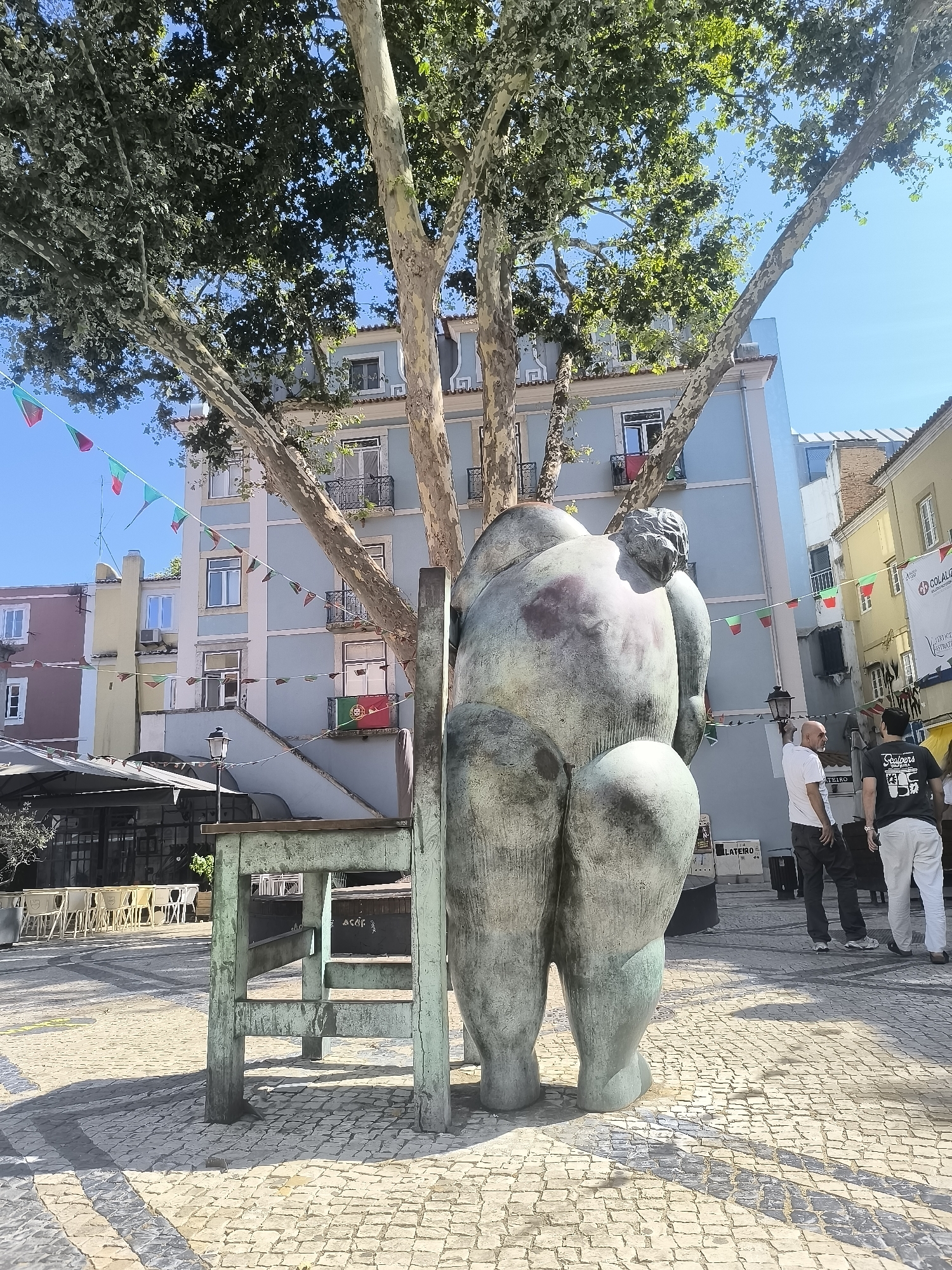 The image captures a public square in Setúbal, Portugal, with a large, weathered bronze statue of a human figure dominating the foreground. The statue is positioned behind a rustic wooden structure, possibly a stage or seating area, with weathered, green-painted legs and a wooden top. The statue's back is to the viewer, showcasing its voluminous form.
In the background, a light blue building with white trim and windows with balconies, some adorned with Portuguese flags, stands under a bright blue sky. Several trees with distinctive mottled bark and lush green foliage partially obscure the building. A string of small, colorful triangular flags, some bearing Portuguese colors, stretches across the scene, suggesting a festive occasion.
To the right of the statue, two men are walking away from the camera. One man, wearing a white t-shirt and dark pants, is partially visible. The other, in a black t-shirt with white text and light-colored pants, is more clearly seen. Further to the right, part of a building with a yellow facade is visible, with a banner that has some text on it. The ground of the square is paved with light-colored cobblestones, creating a textured surface with distinct shadows from the overhead trees and the statue. Outdoor seating with white chairs and tables is visible in the middle ground, to the left of the statue, suggesting an area for cafes or restaurants. A street lamp is also present.