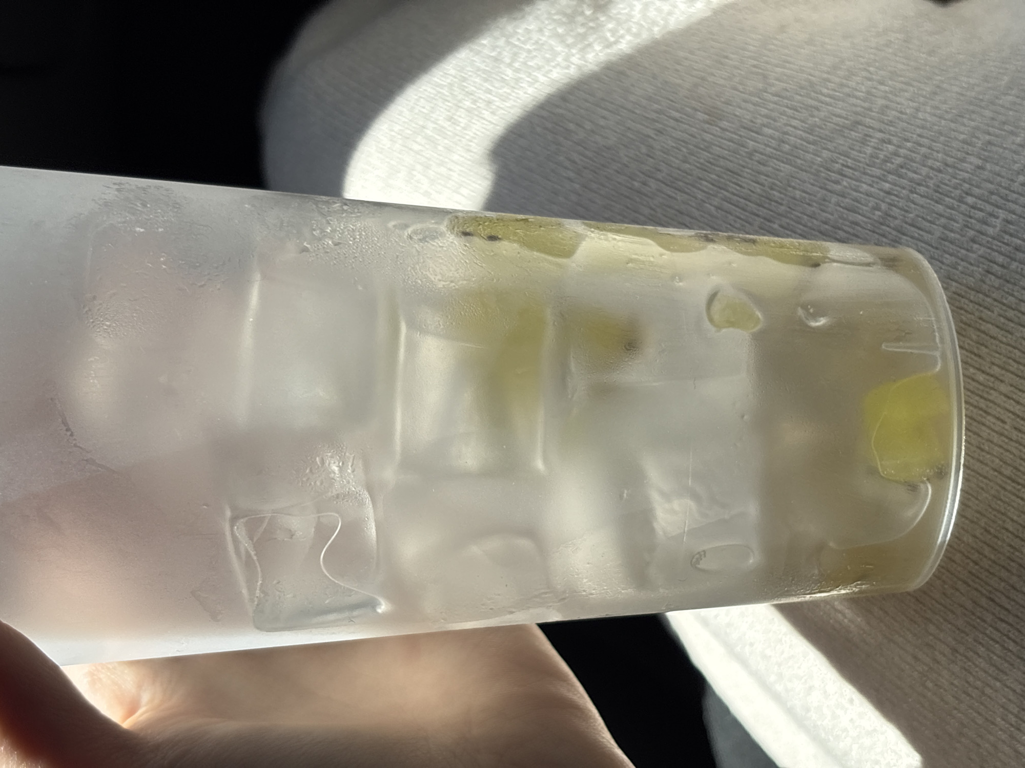 A person's hand holds a frosted plastic cup filled with ice cubes and what appear to be slices of kiwi. The cup is condensation-covered, suggesting it contains a cold beverage. The background is dark, with a white, textured surface visible on the right, possibly a piece of clothing or upholstery. The scene is illuminated by sunlight, casting shadows. The location is stated to be Saint Petersburg, Russia, although there are no visible landmarks or contextual clues within the image to support this.